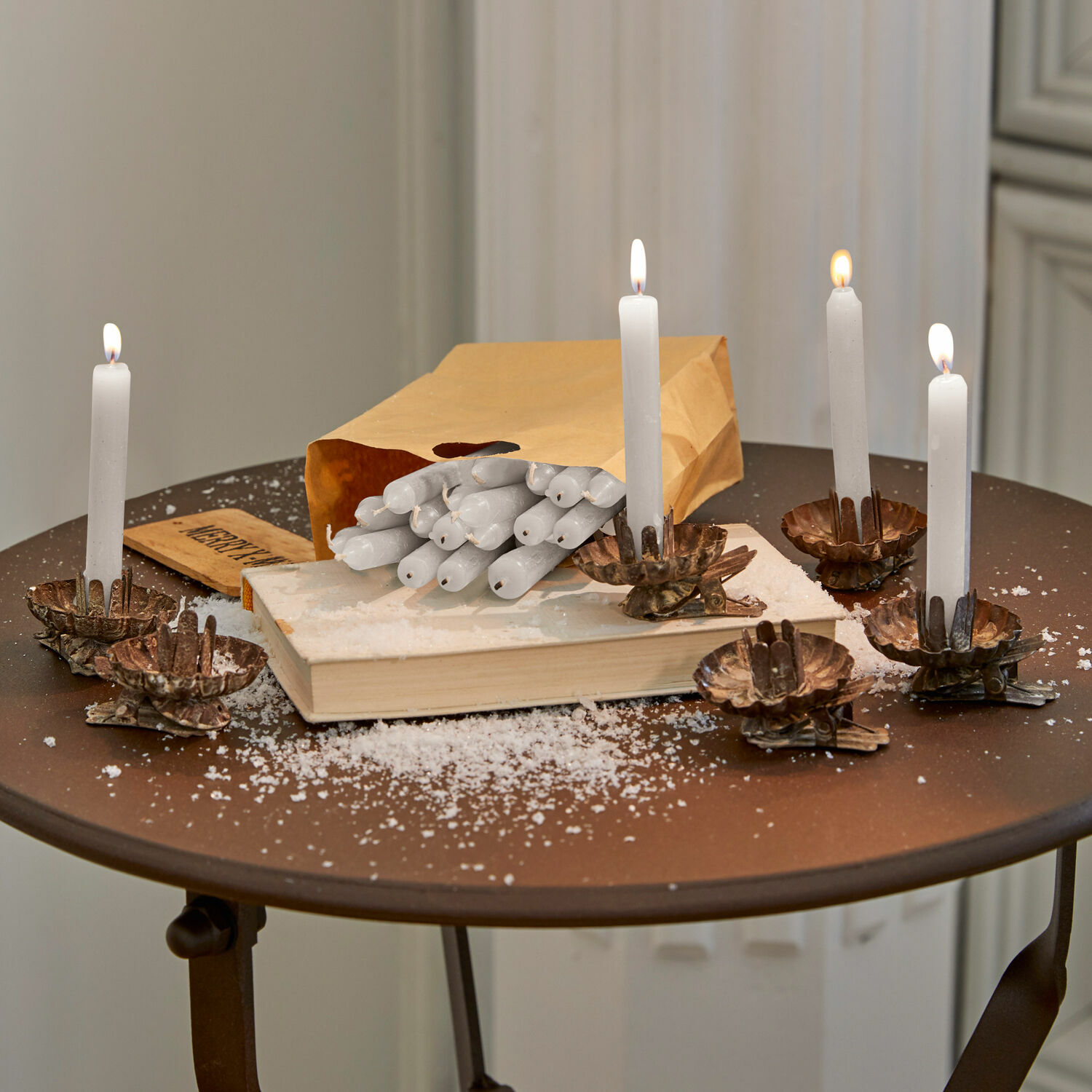 Lot de 20 bougies Noufa Bougies blanches en cire sur des chandeliers en métal cuivré sur une table brune avec sac en papier