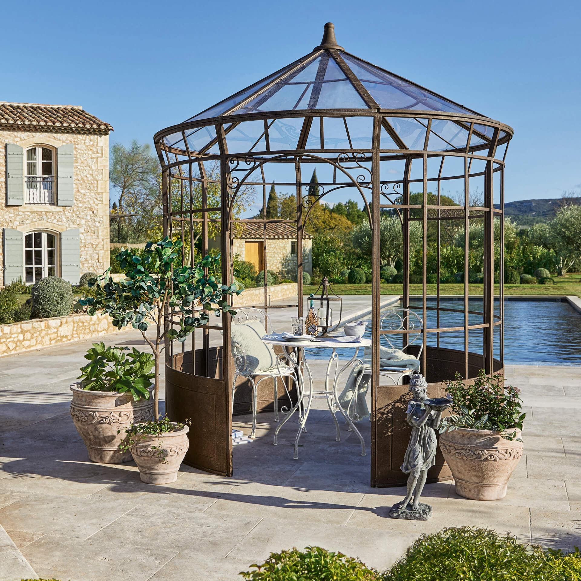 Jardin avec une gloriette en métal rouillé, table et chaises blanches, entourée de pots de plantes
