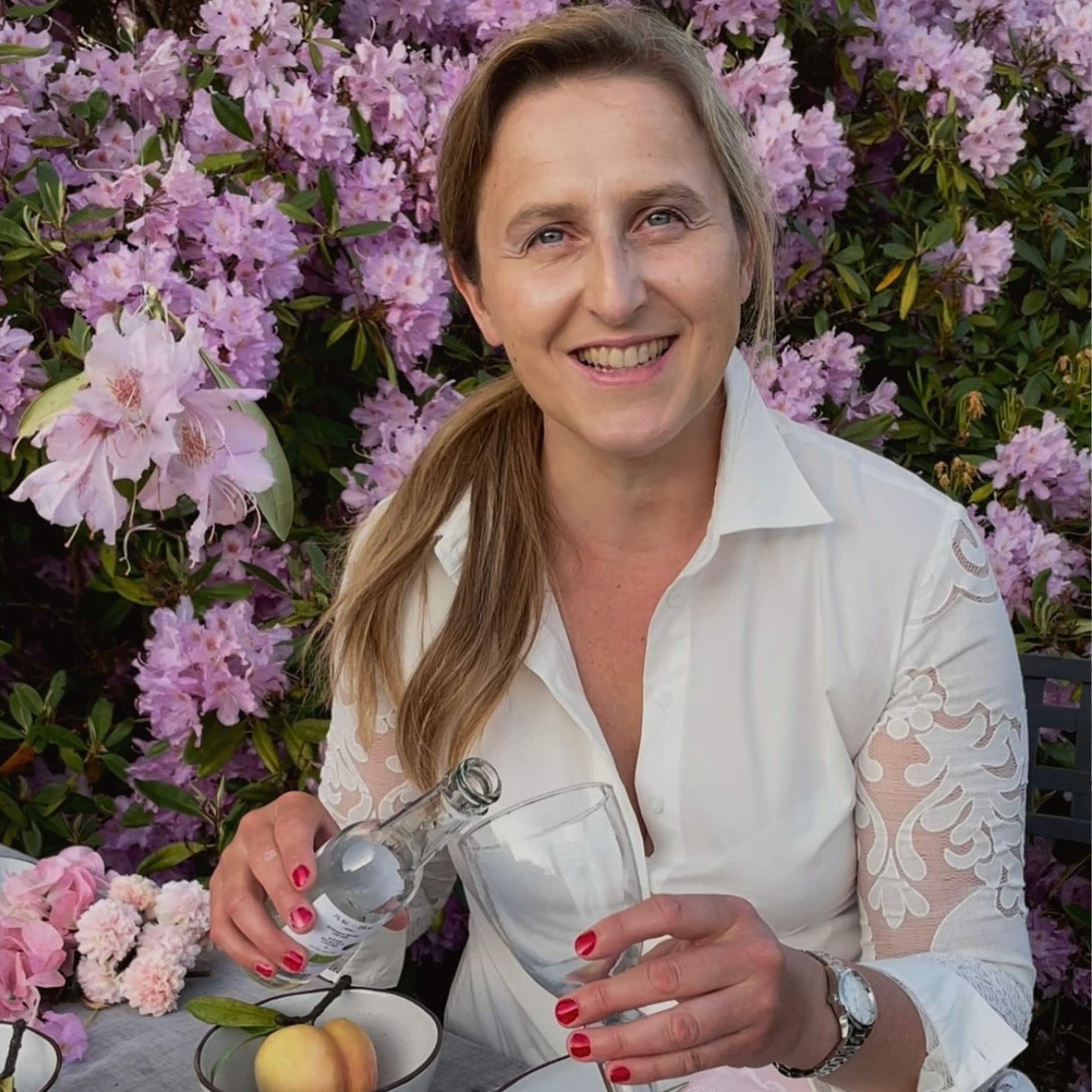 Femme en chemise blanche à manches en dentelle versant une boisson dans un verre devant des fleurs roses