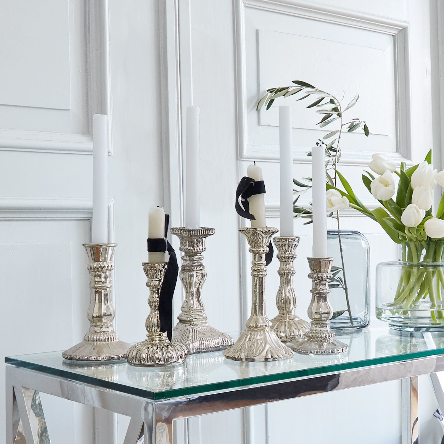 Lot de 6 chandeliers Adolpha Ensemble de chandeliers en métal argenté avec bougies blanches sur une table en verre dans un intérieur lumineux