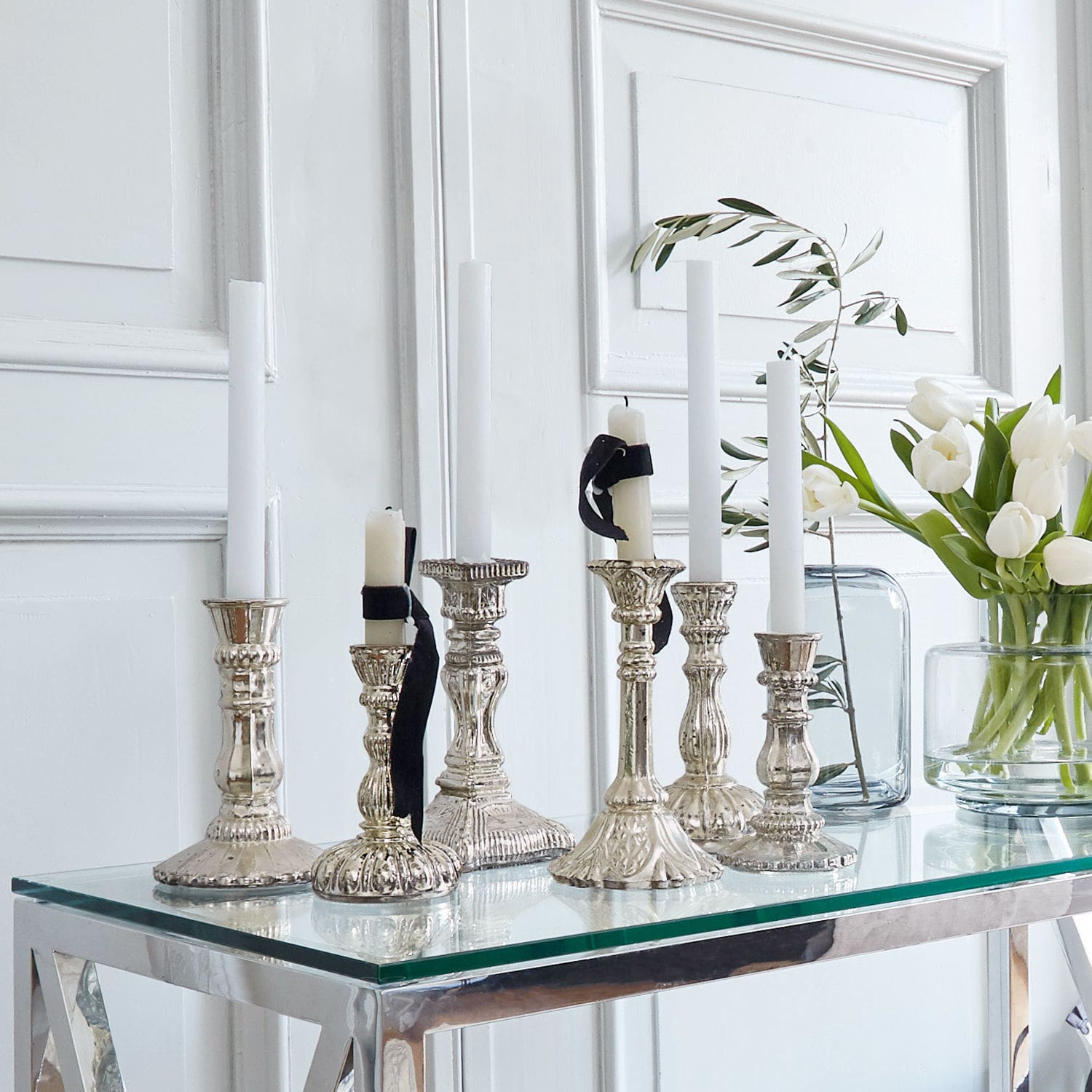 Lot de 6 chandeliers Adolpha Ensemble de chandeliers en métal argenté avec bougies blanches sur une table en verre dans un intérieur lumineux
