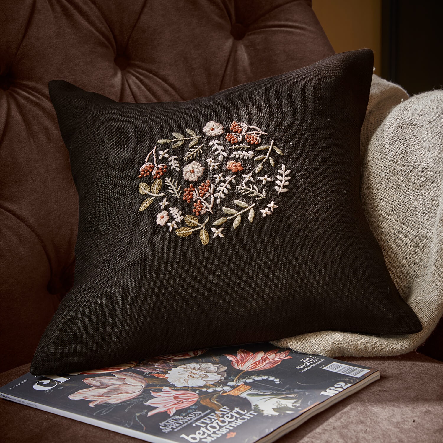 Coussin noir en lin avec broderie florale pastel posé sur un fauteuil avec magazine