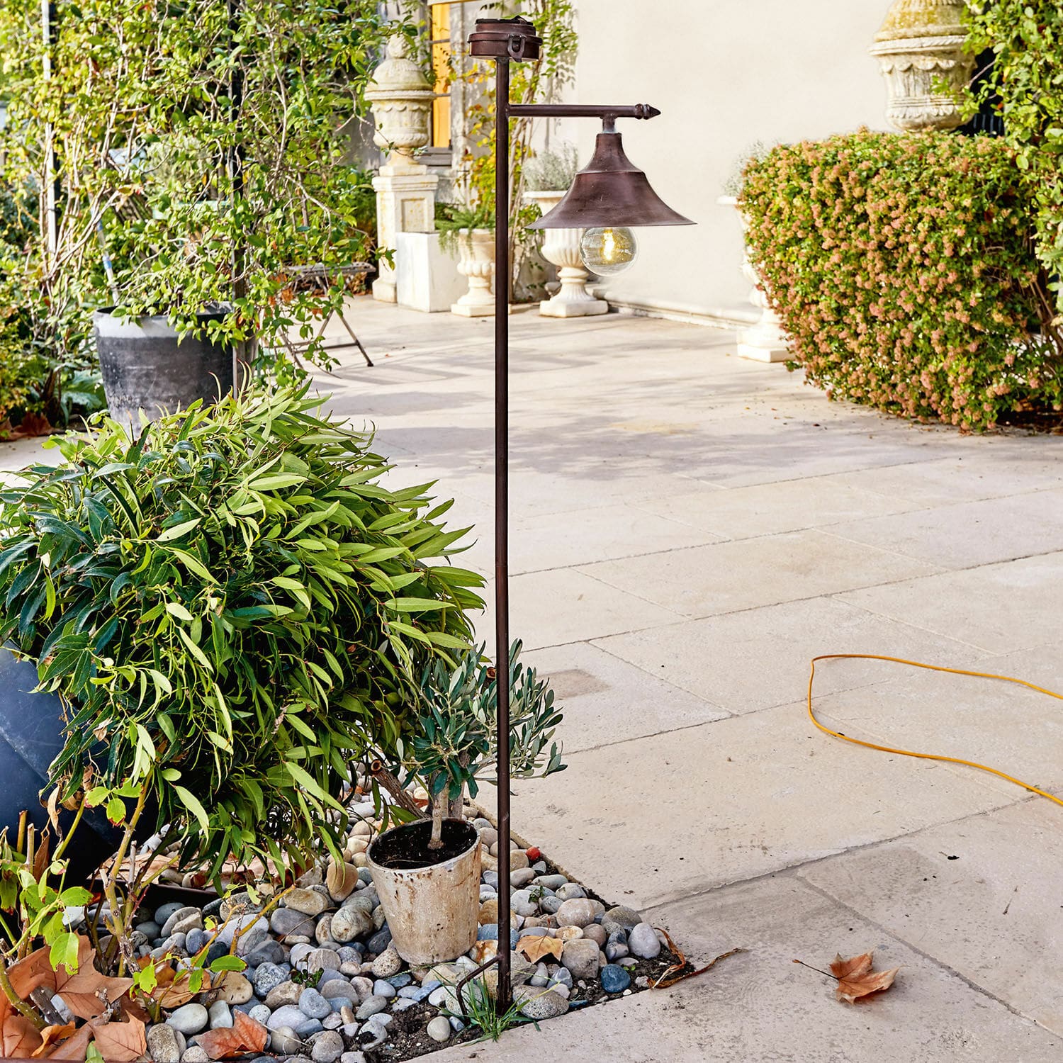 Lanterne solaire Kaley Lampe de jardin en métal couleur rouille avec ampoule, posée sur des pierres parmi des plantes vertes