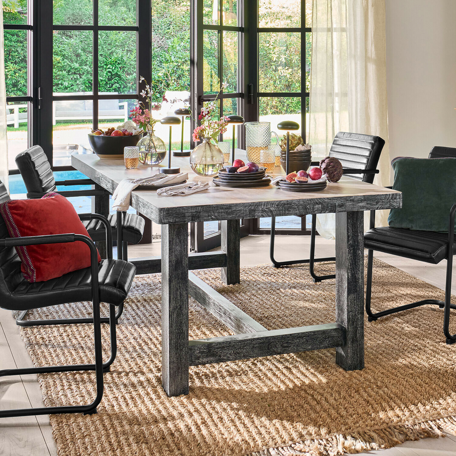 Table Oaklynne Table à manger en bois gris avec chaises noires et décorations, devant de grandes fenêtres lumineuses