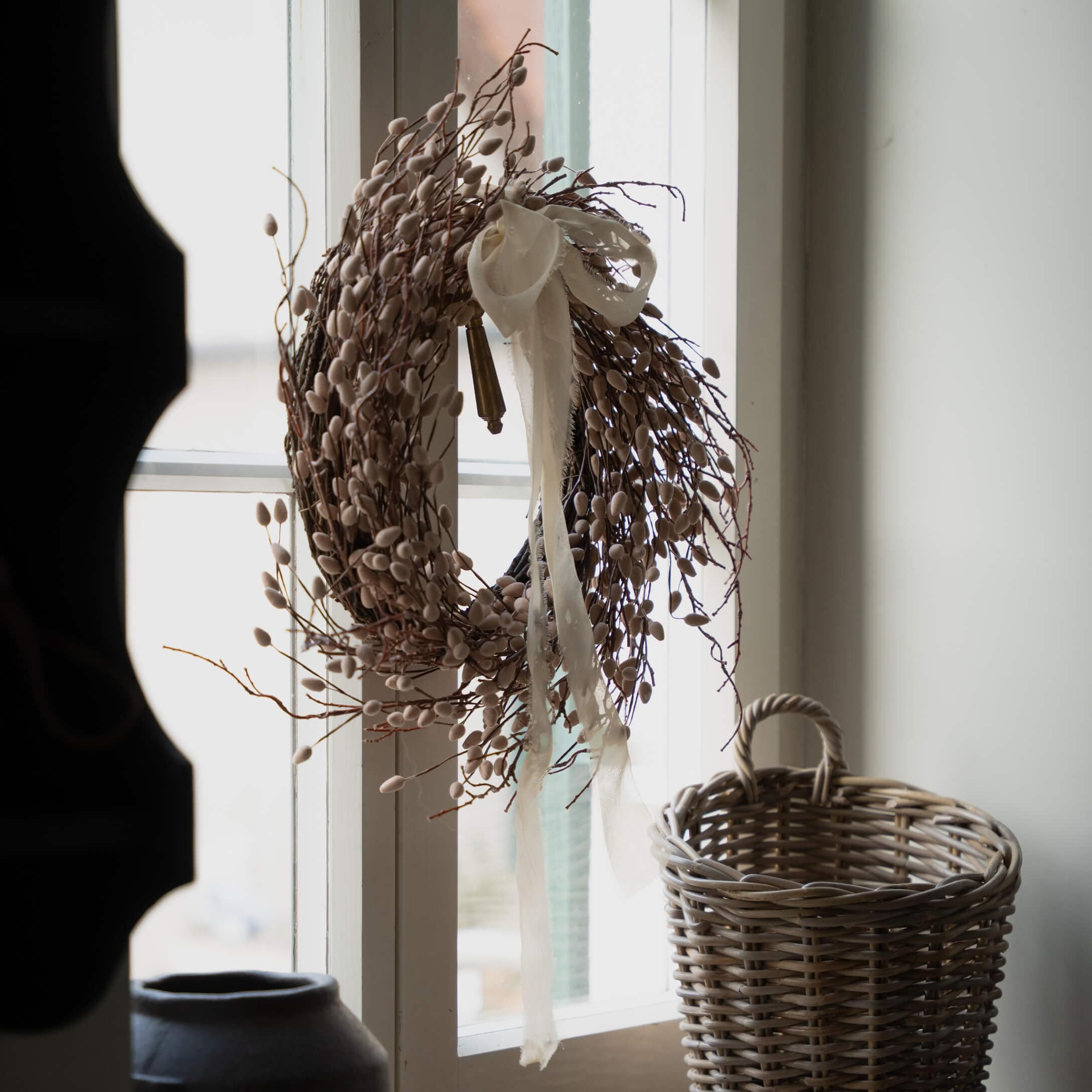 Couronne décorative beige en branches et bourgeons avec ruban blanc suspendue à la fenêtre, panier en osier à côté