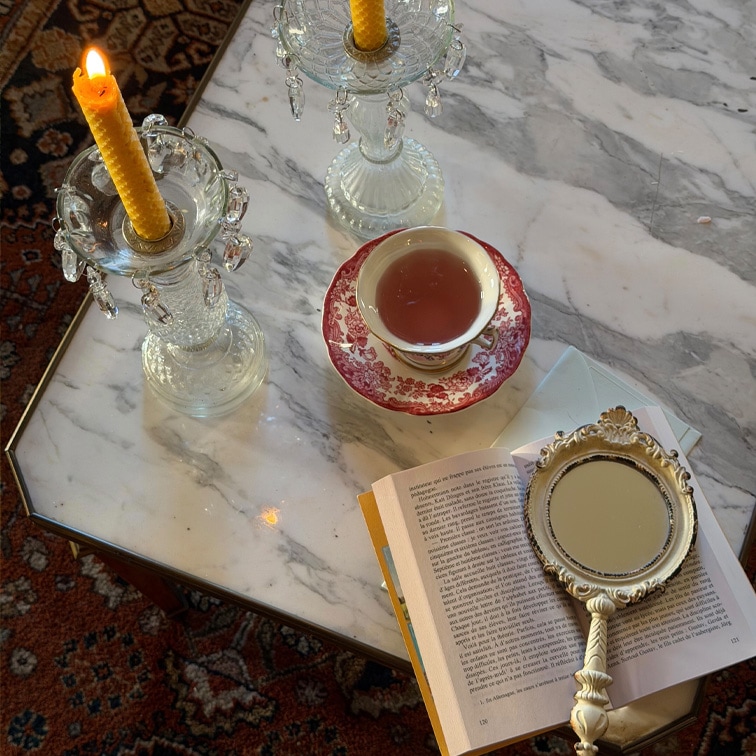 Bougeoirs en verre avec bougies jaunes, tasse à motif rouge sur table en marbre avec livre ouvert et miroir à main