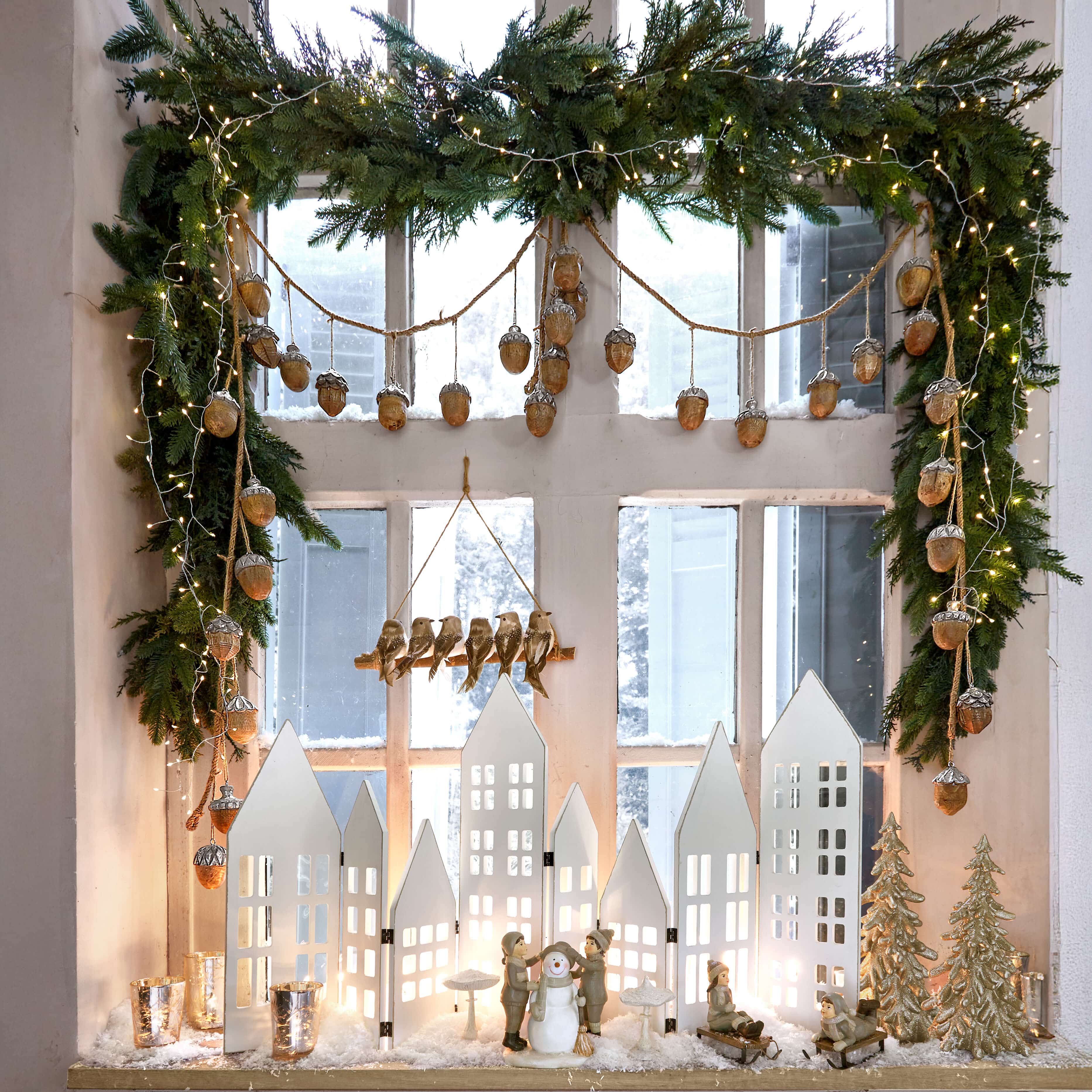 Décoration de Noël avec maisons blanches, figurines d'enfants et bonhomme de neige sur rebord avec guirlande verte et lumières