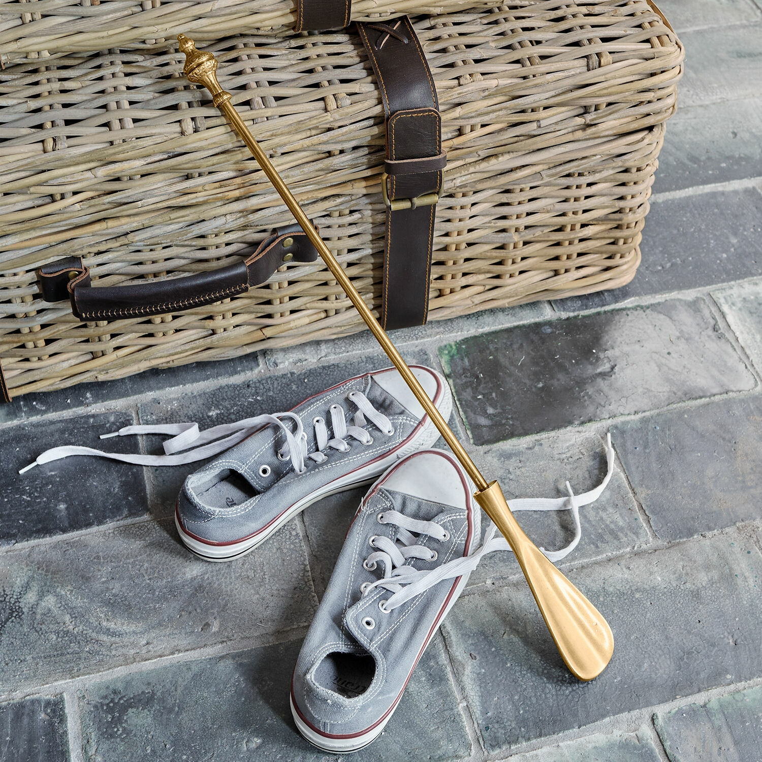 Chausse-pied Orveline Chausse-pied en métal doré posé sur un sol en pierre avec un panier en osier et des baskets grises