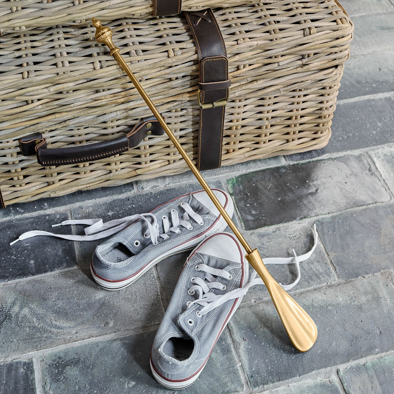 Chausse-pied Orveline Chausse-pied en métal doré posé sur un sol en pierre avec un panier en osier et des baskets grises