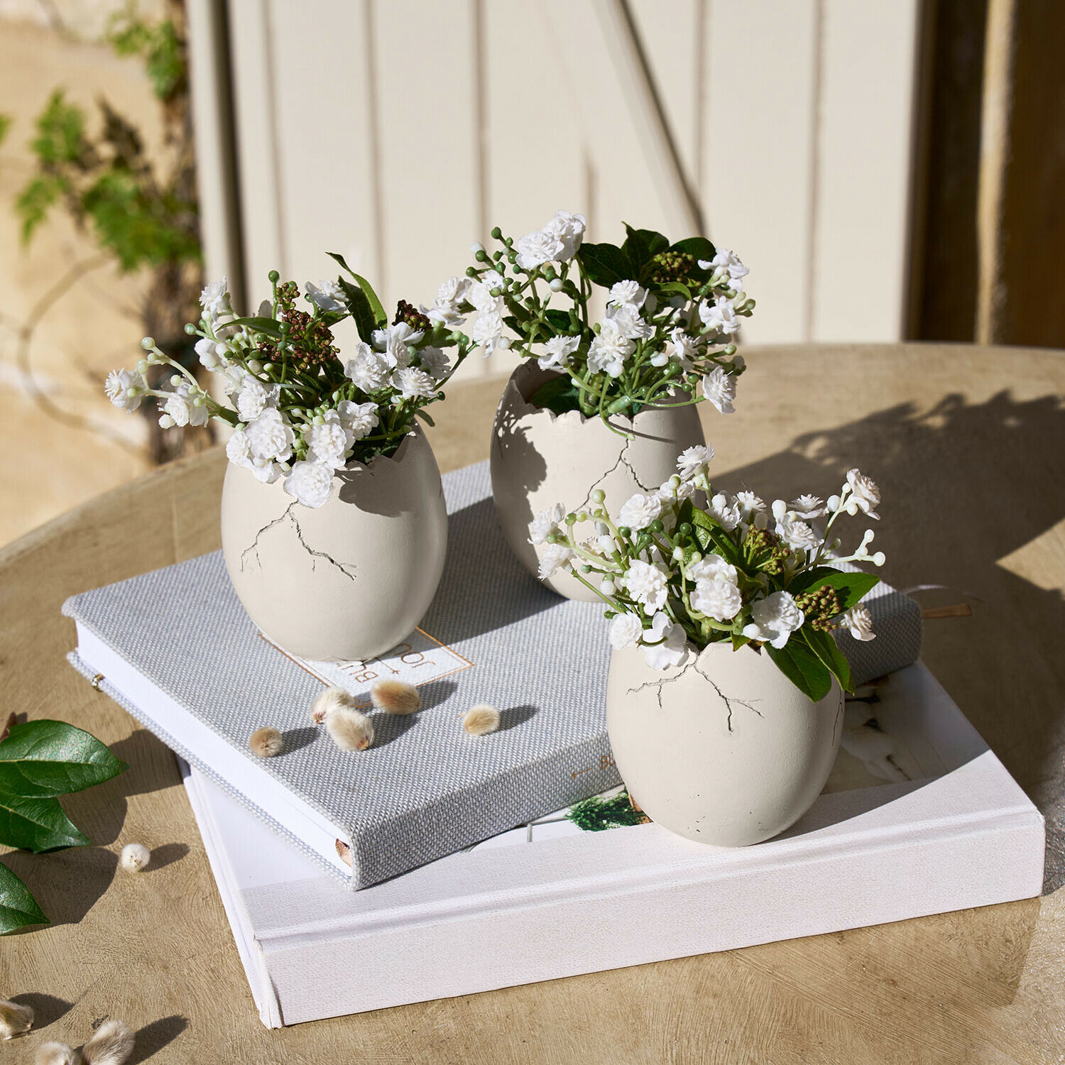 Lot de 3 œufs décoratifs Waverlye Trois vases en céramique blanche en forme d'œufs avec des fleurs blanches sur une table en bois avec des livres