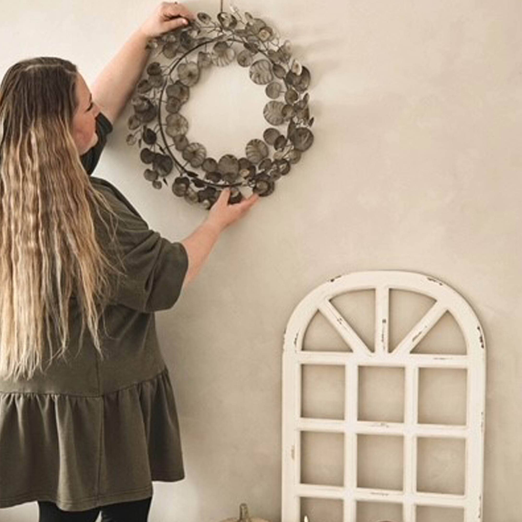 Femme accrochant une couronne décorative en métal brun sur un mur clair dans un style rustique