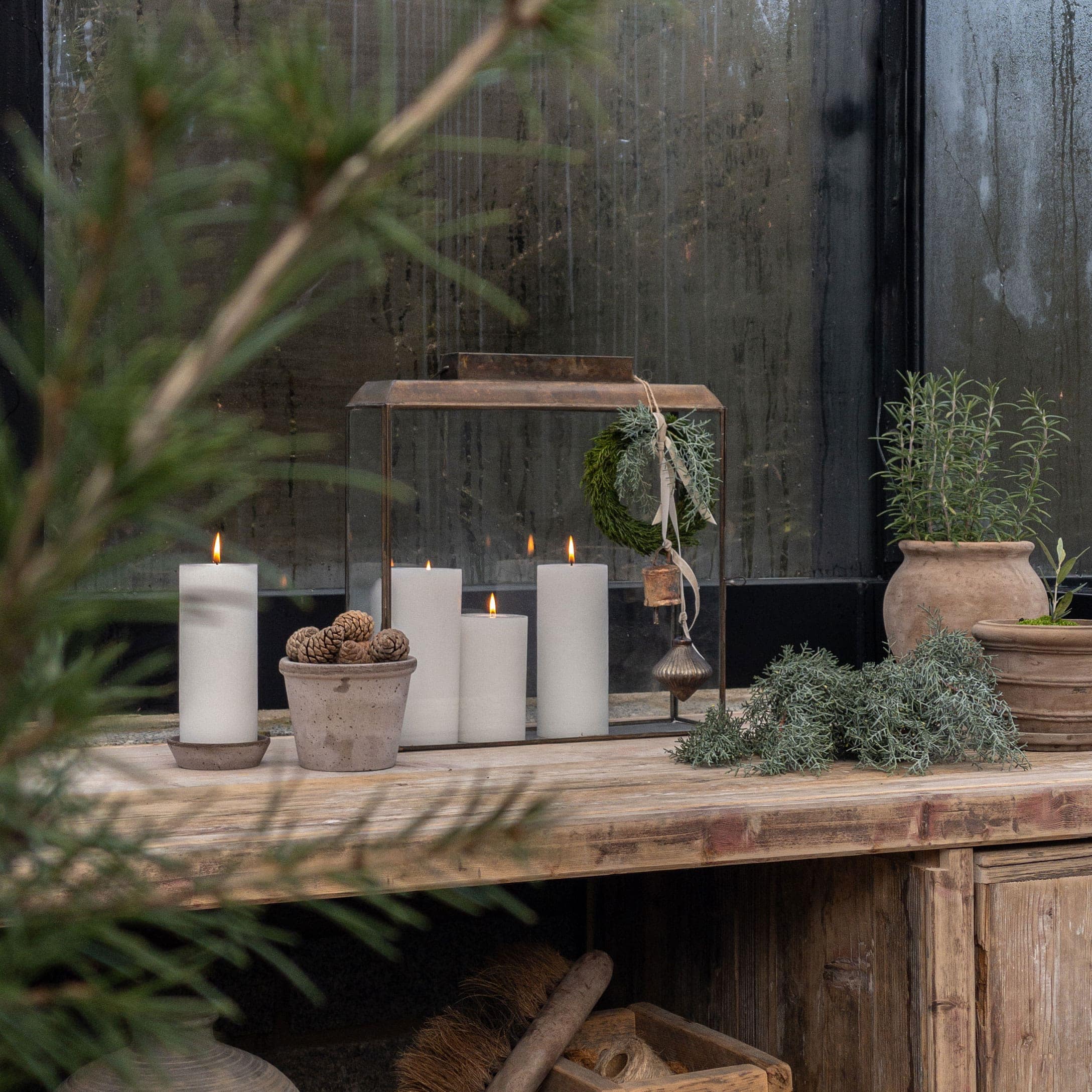 Table en bois rustique avec bougies blanches et pots de plantes vertes devant une vitre embuée