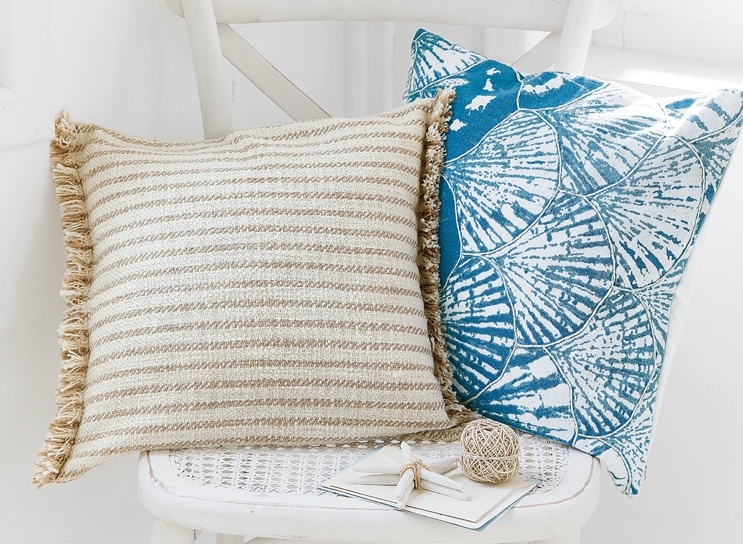 Deux coussins décoratifs sur une chaise blanche, un beige rayé avec franges, l'autre bleu à motif coquillage
