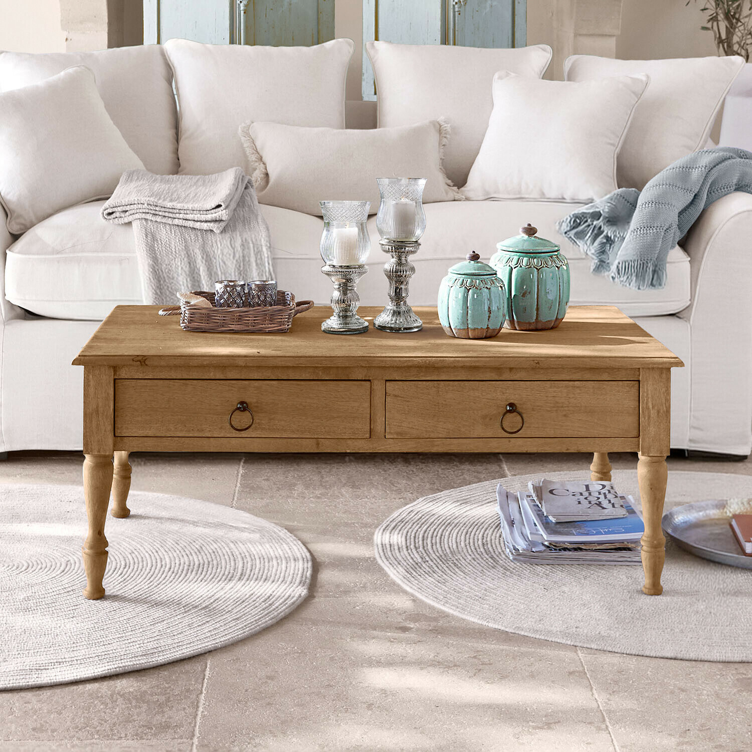 Table basse en bois avec tiroirs devant un canapé clair, décorations et plaids posés dessus