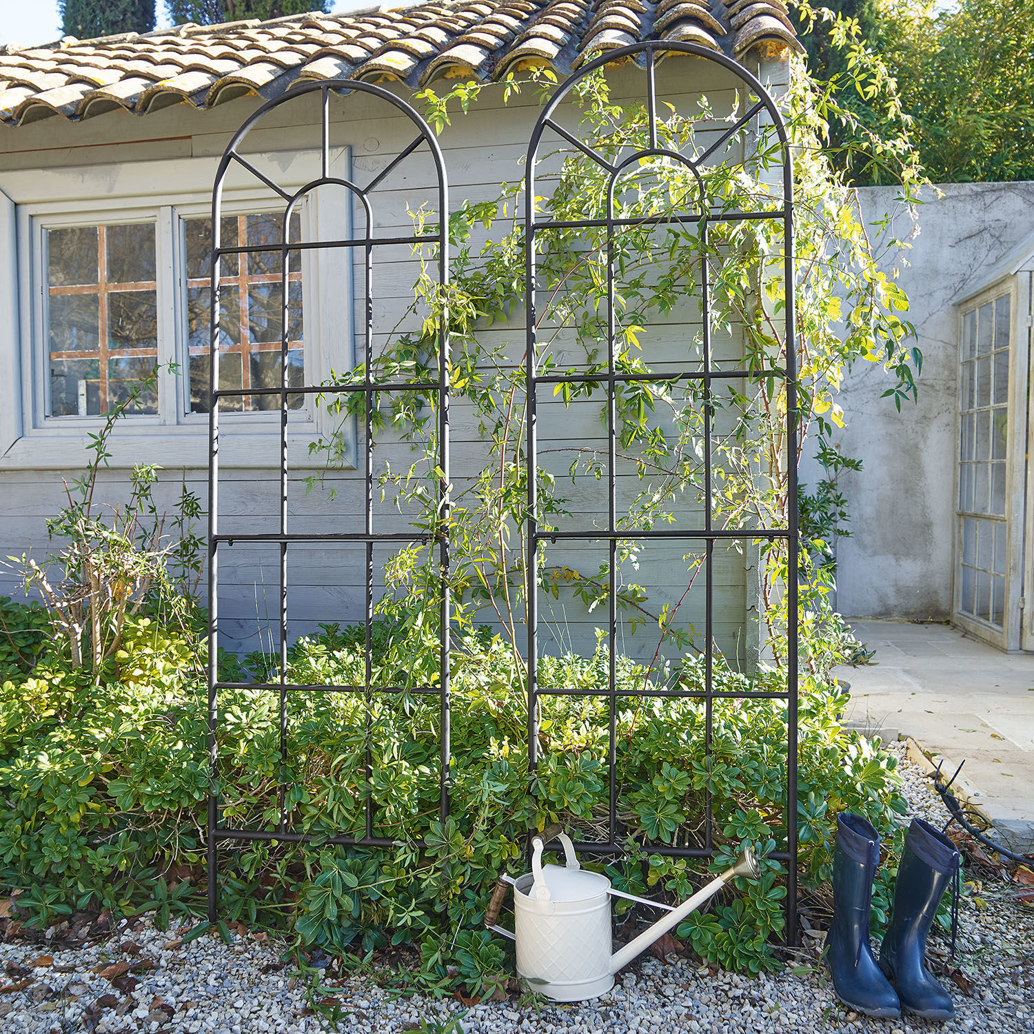 Lot de 2 treillis Ealdred Treillis de jardin en métal noir avec motif arrondi devant des plantes et une maison, arrosoir et bottes bleues
