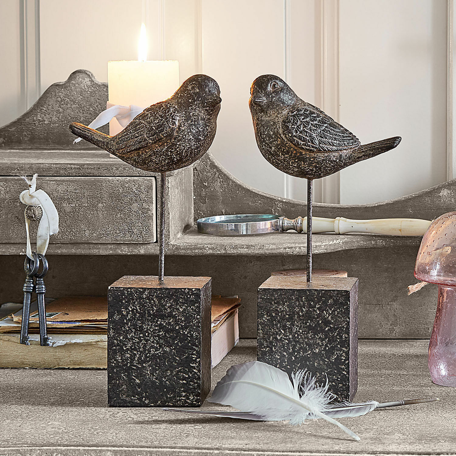 Lot de 2 oiseaux décoratifs Juxivel Oiseaux décoratifs en métal foncé sur socles carrés posés sur une table rustique avec une plume