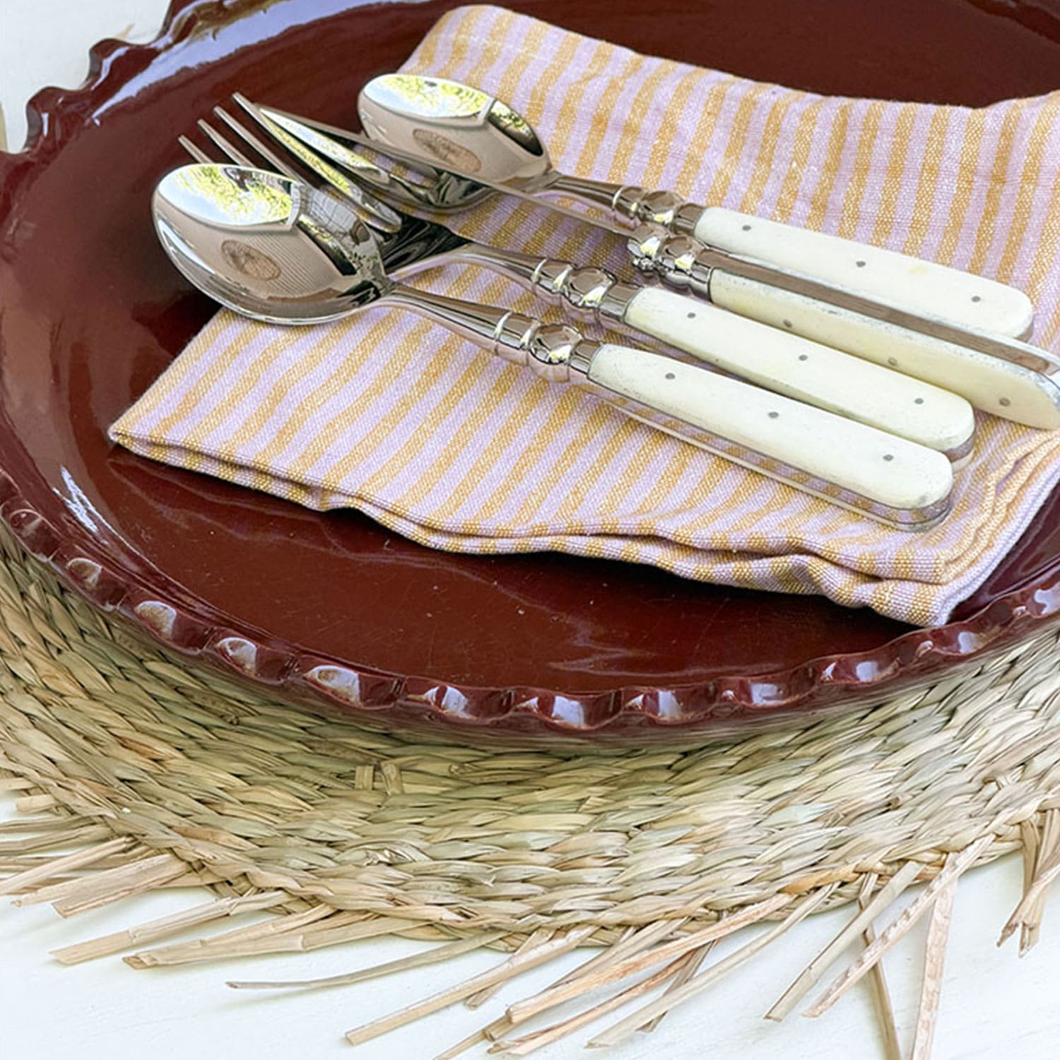 Ensemble de couverts avec manches blancs sur une serviette rayée posée sur une assiette marron et un dessous de plat tressé
