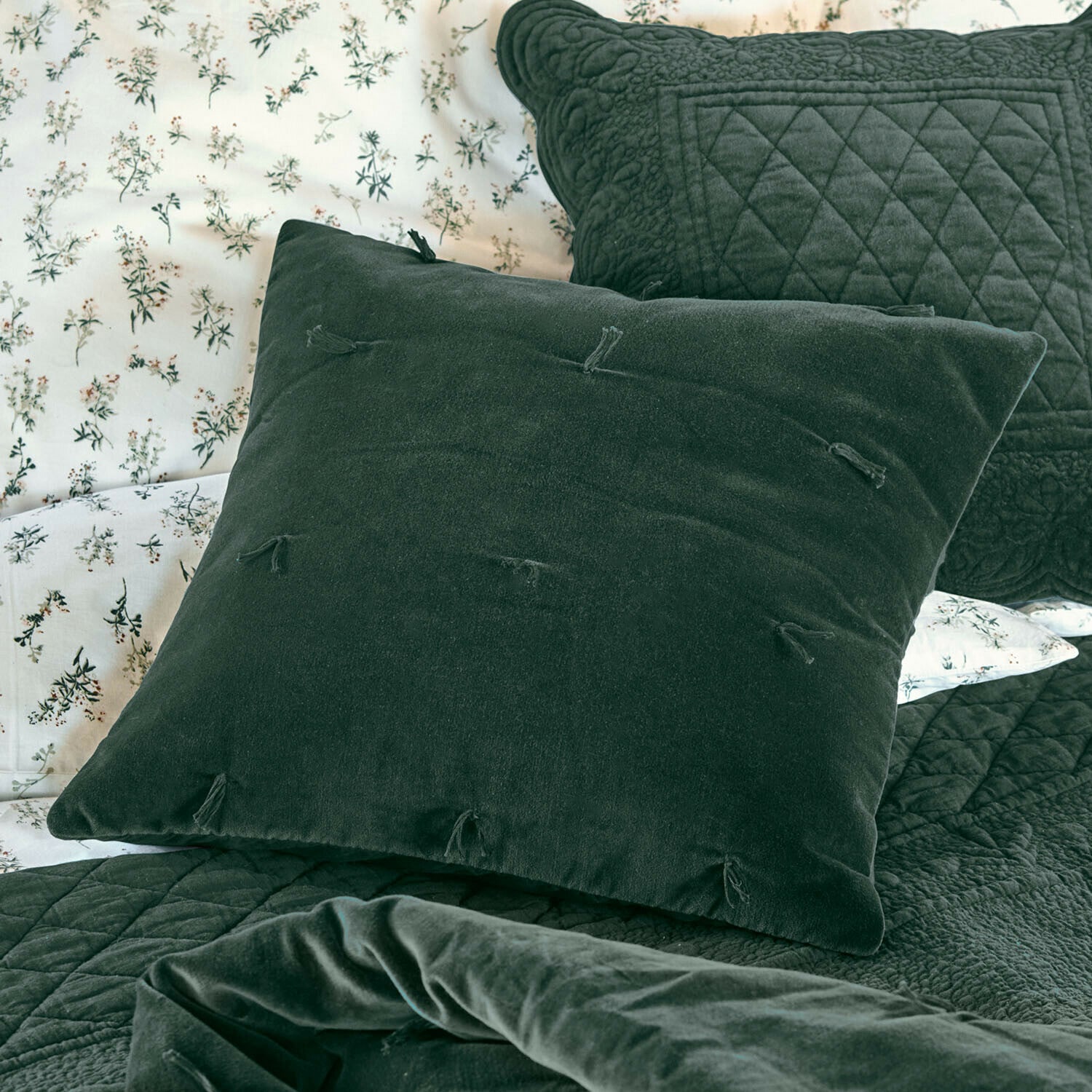 Coussin Orélisse Coussin décoratif vert en velours avec pompons sur lit avec couvre-lit matelassé et draps à fleurs
