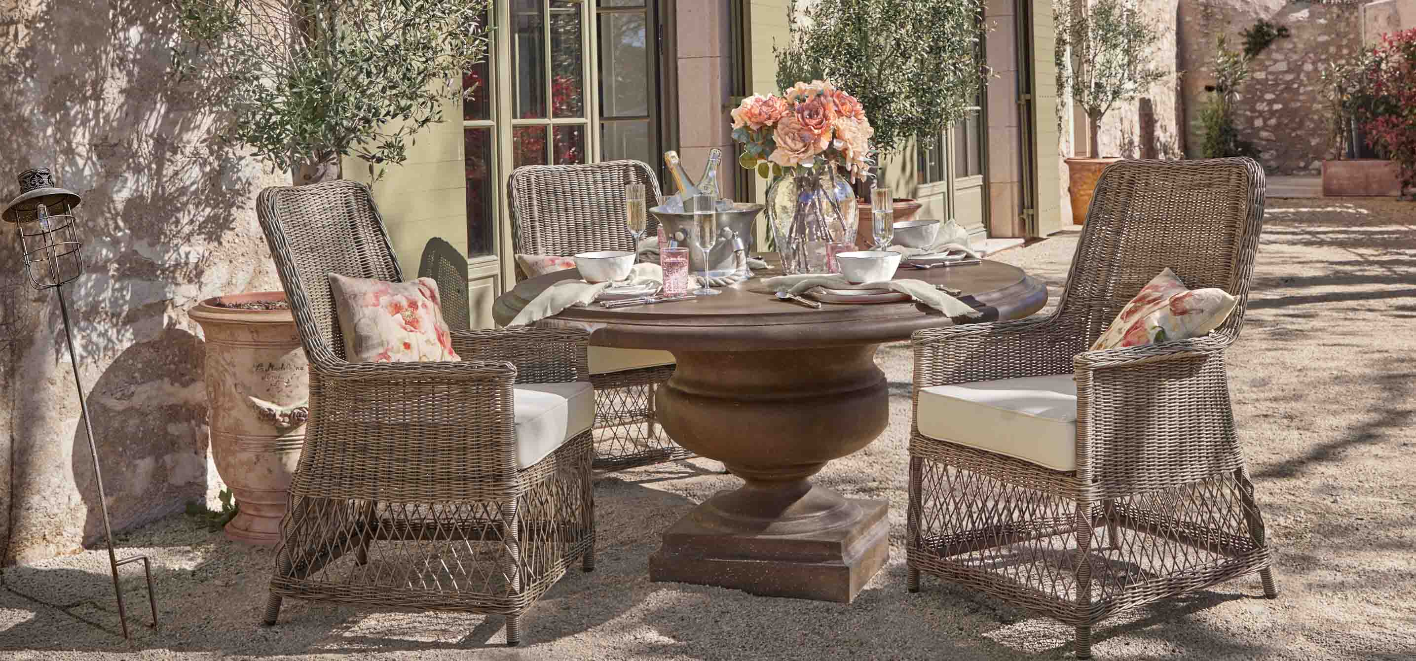 Table ronde en métal brun avec quatre chaises en osier et coussins clairs dans un jardin ensoleillé