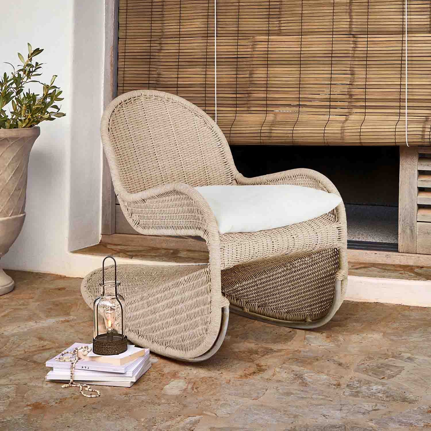 Fauteuil en osier beige avec coussin blanc sur terrasse, lanterne et livres posés sur sol en pierre
