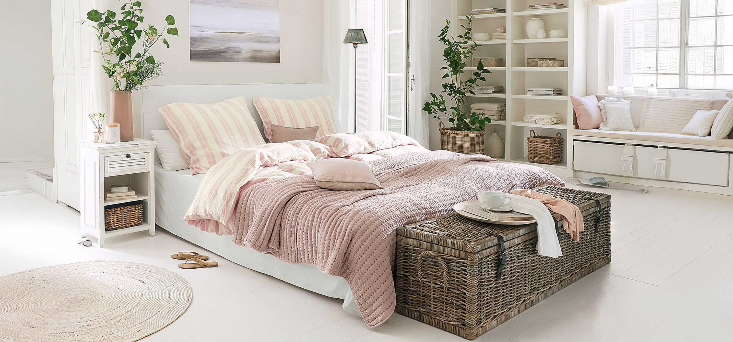 Parure de lit douce rose et beige dans une chambre lumineuse avec coffre en osier au pied du lit