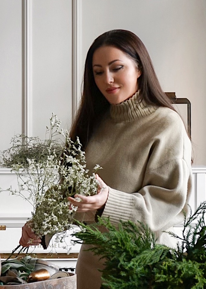 Femme en pull beige à col roulé tenant des fleurs blanches délicates dans un intérieur lumineux