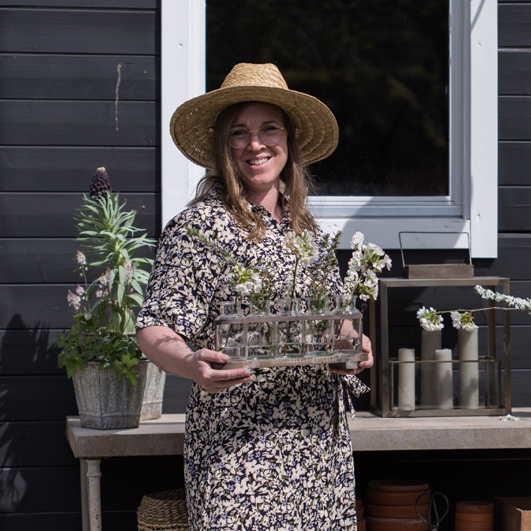 Femme avec chapeau tenant un vase en verre avec des fleurs devant un mur noir et une fenêtre