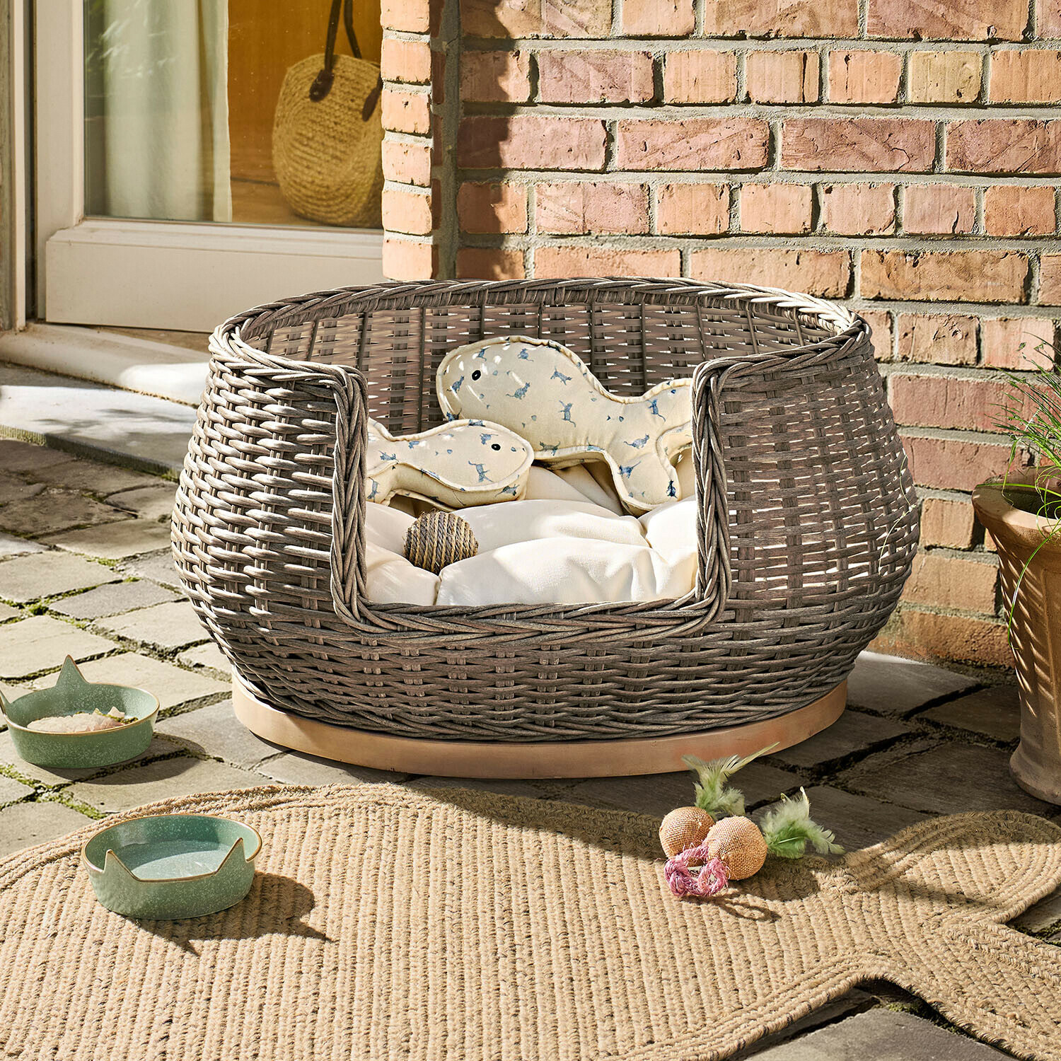 Panier rond en osier gris avec coussin crème et jouets pour chien sur terrasse en pierre