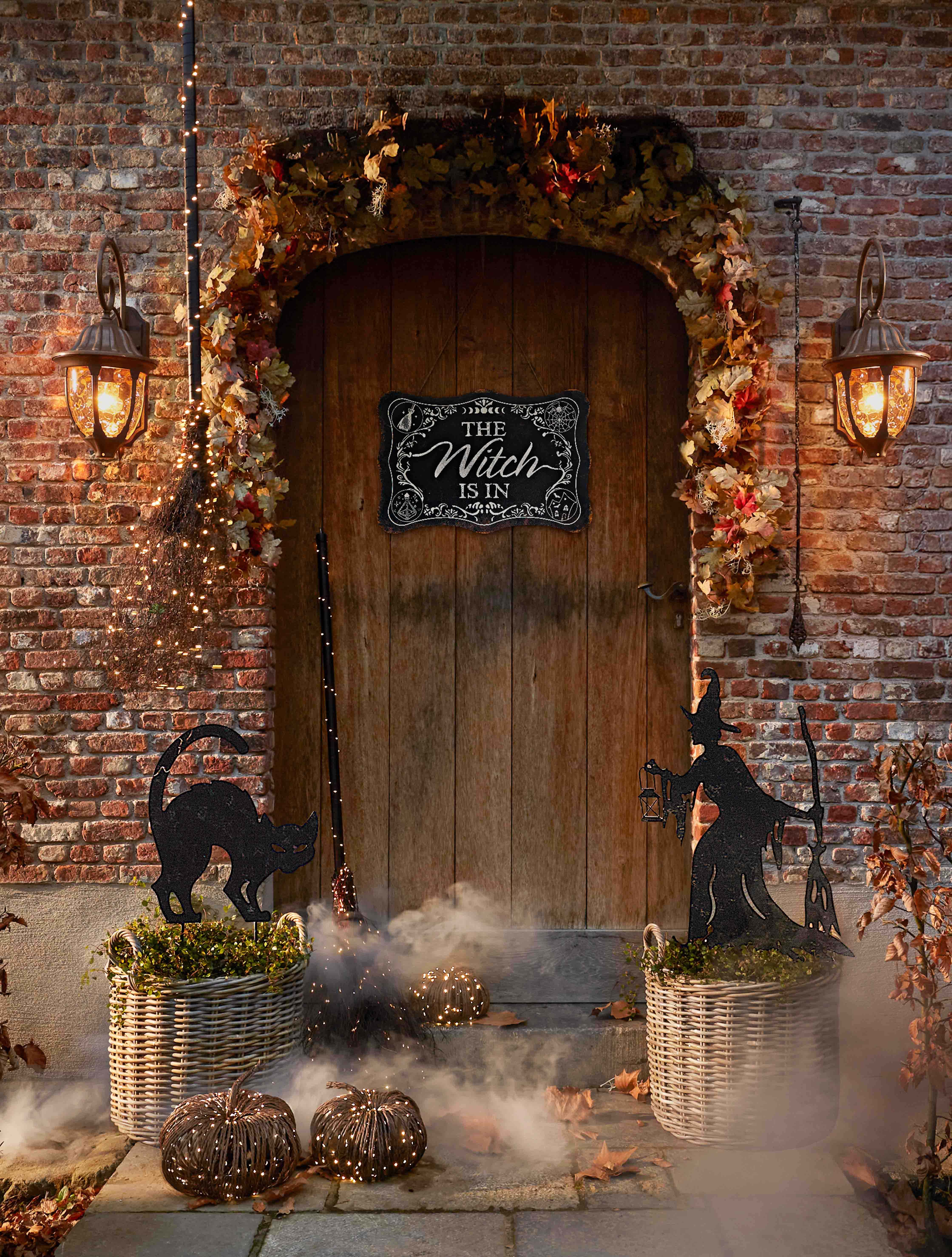 Décoration d'Halloween avec sorcière, chat noir et lanternes devant une porte en bois et mur en briques
