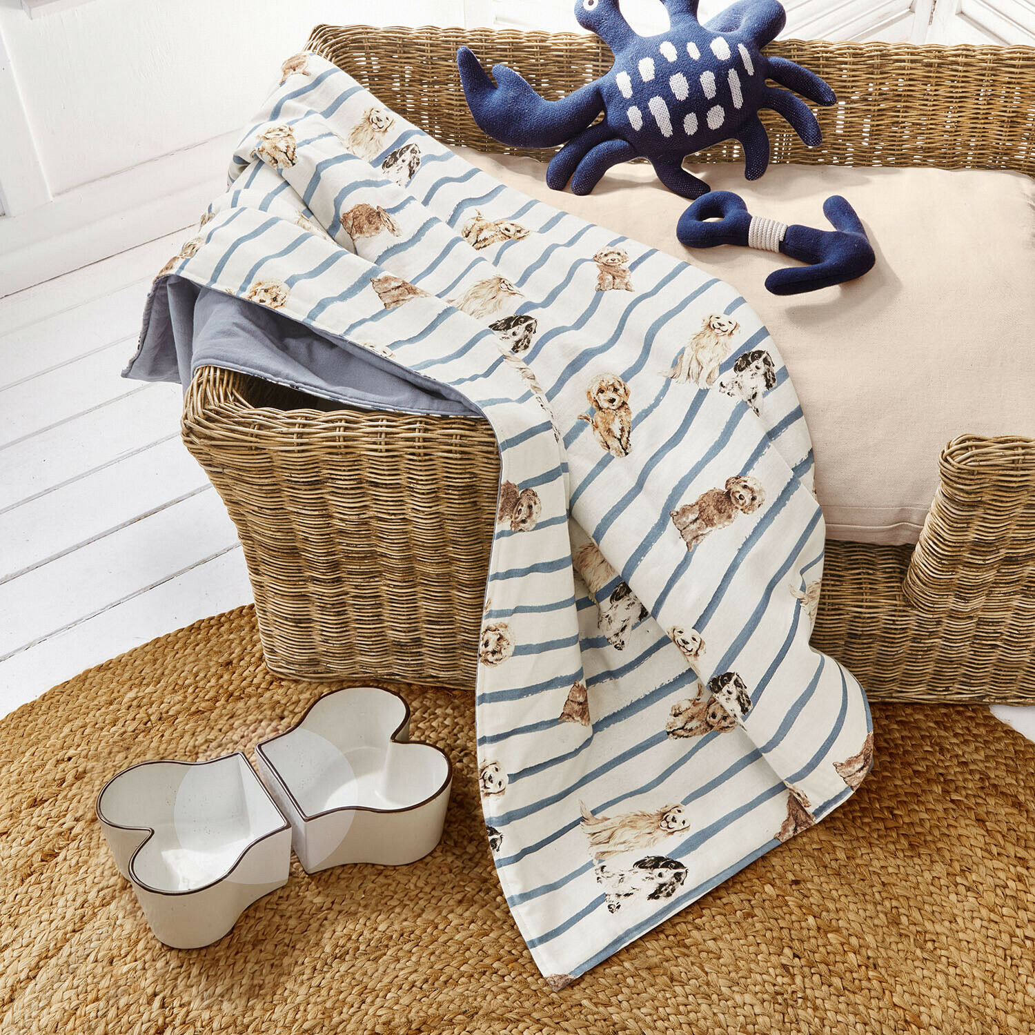 Couverture pour chien rayée bleu et blanc avec motifs de chiens sur panier en osier avec jouets et gamelle