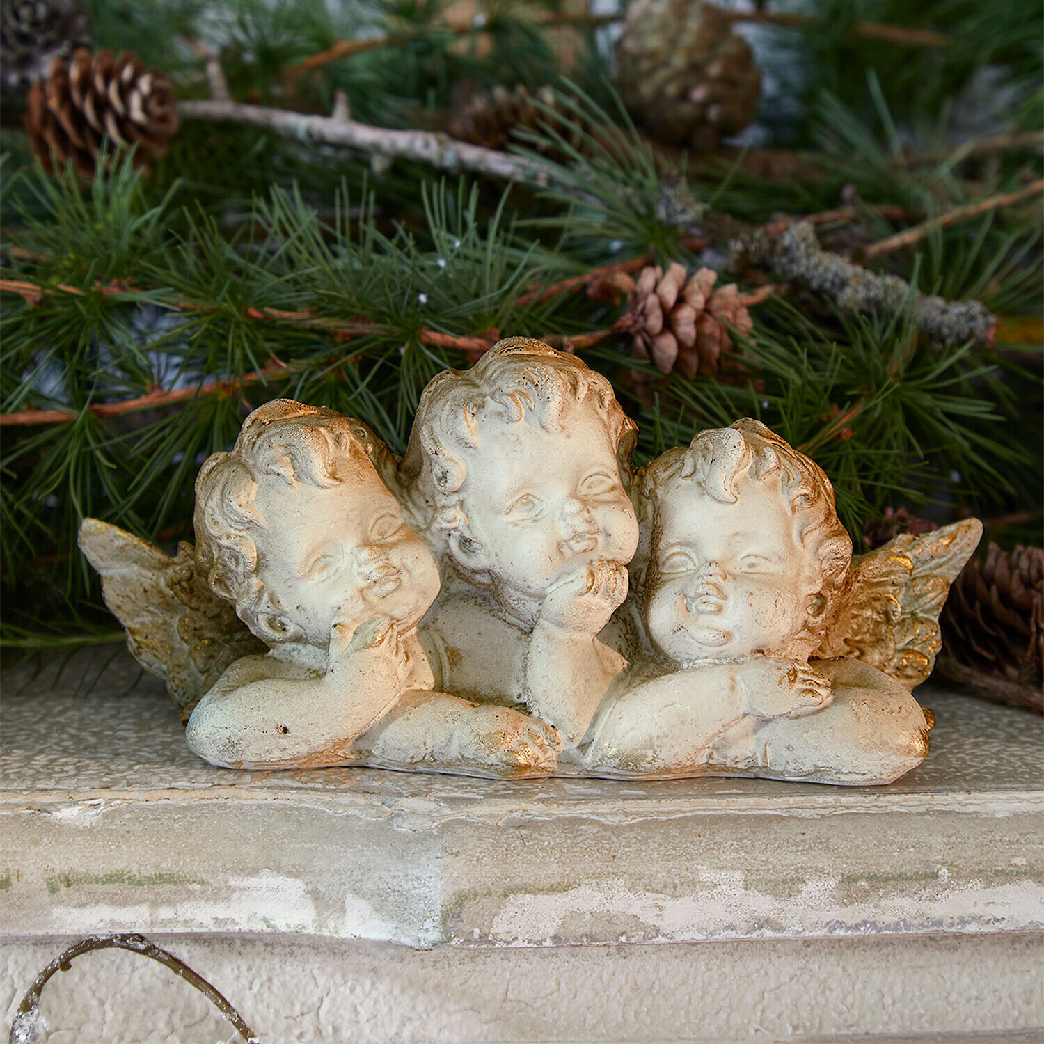 Anges décoratifs Nivardo Figurine de trois anges en pierre claire sur fond de branches de pin vertes, décoration festive