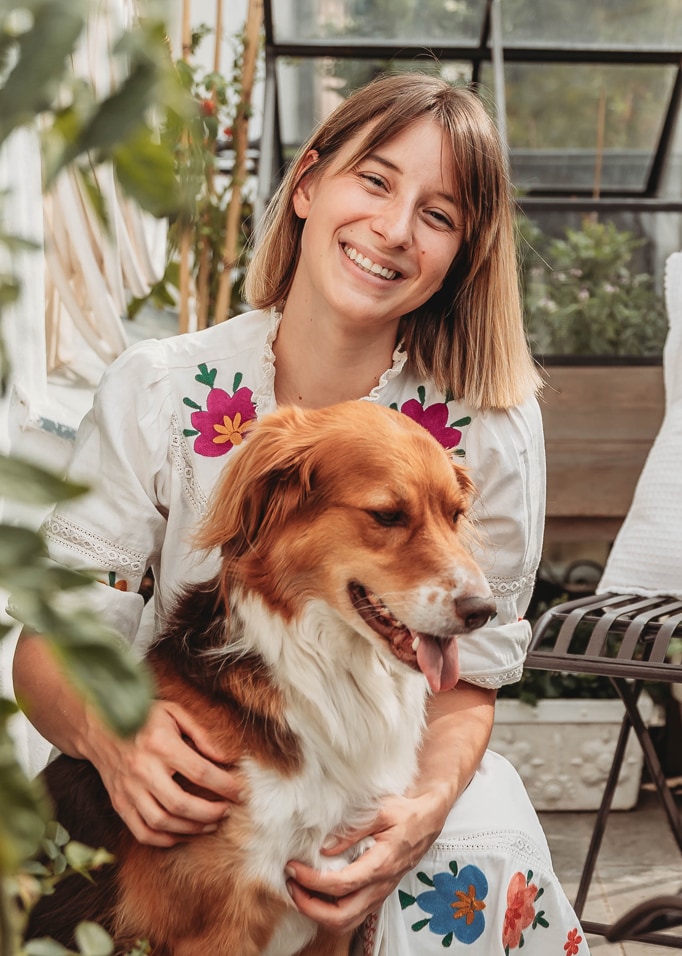 Femme en robe blanche brodée colorée tenant un chien brun et blanc dans un intérieur lumineux avec des plantes