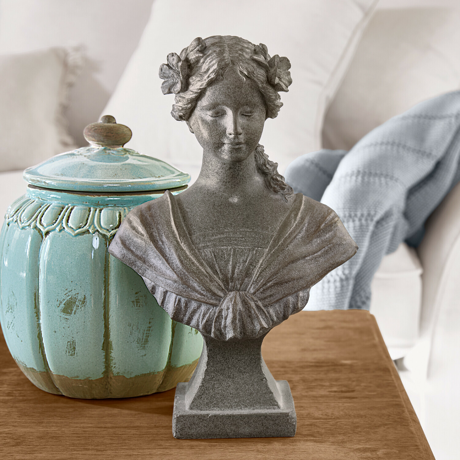 Buste Alexandra Buste féminin en pierre grise avec fleurs dans les cheveux sur table en bois à côté d’un pot en céramique turquoise
