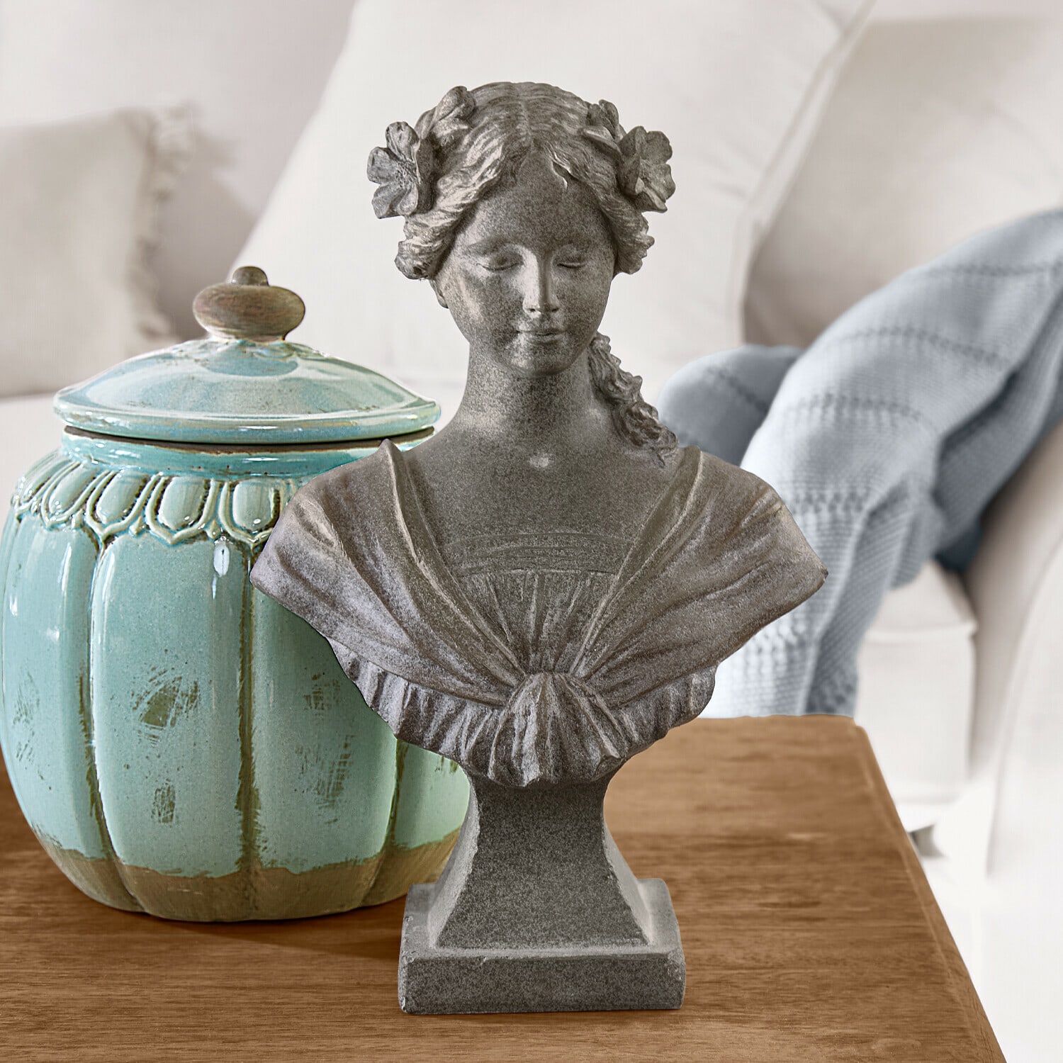 Buste Alexandra Buste féminin en pierre grise avec fleurs dans les cheveux sur table en bois à côté d’un pot en céramique turquoise