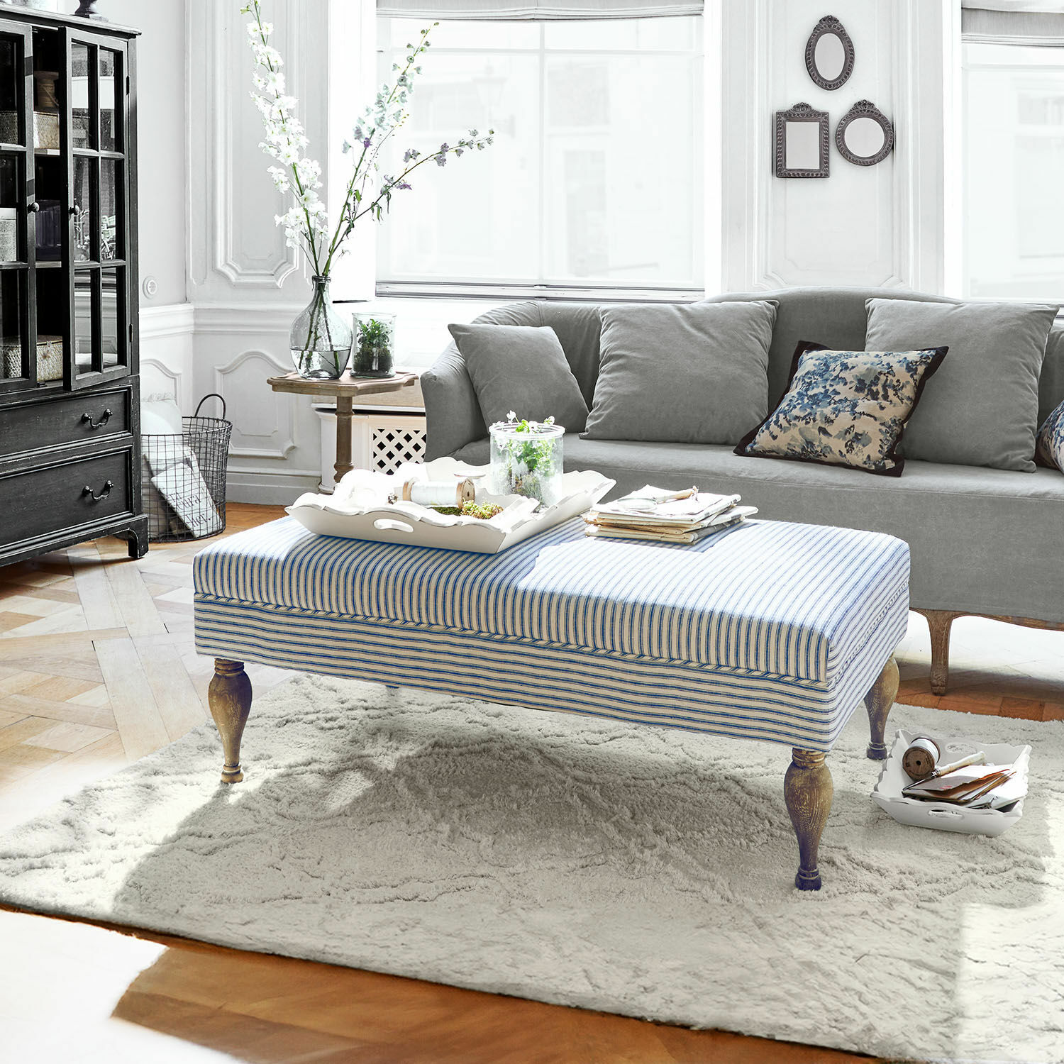 Repose-pieds Ulysette Pouf rayé avec pieds en bois sur tapis clair dans un salon moderne avec canapé gris