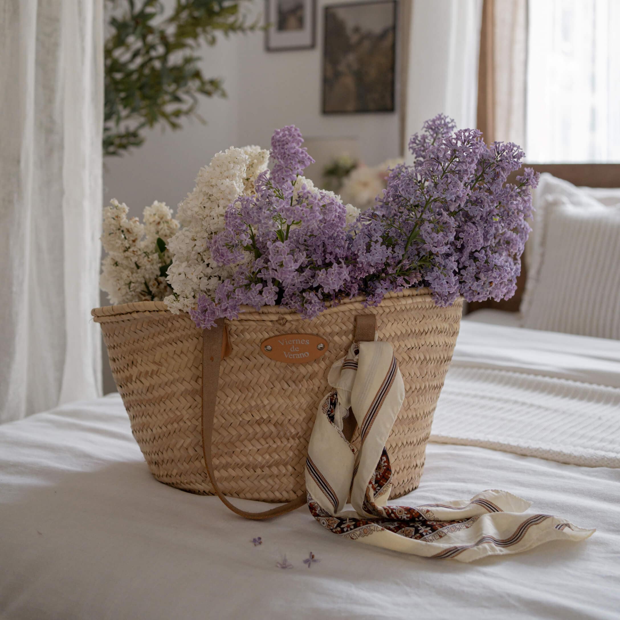 Panier en osier avec fleurs blanches et violettes et foulard posé sur un lit blanc dans une chambre lumineuse
