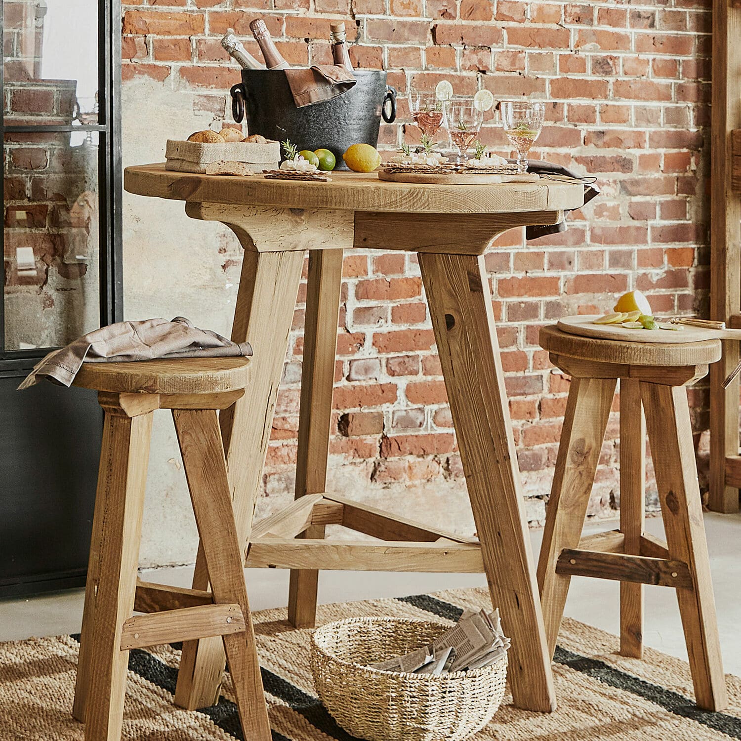 Table haute Stonington Table haute en bois naturel avec deux tabourets assortis devant un mur en briques, style loft