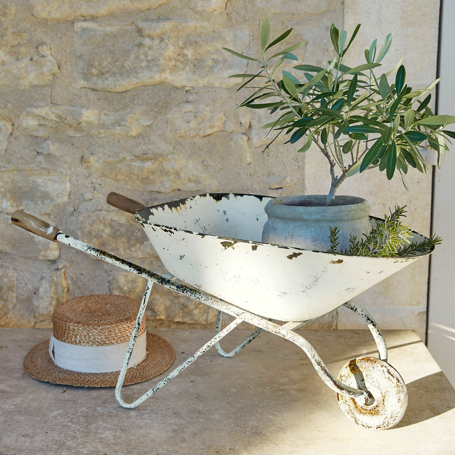 Brouette décorative Padoux Brouette en métal blanc vieilli avec pot et plantes vertes devant un mur en pierre