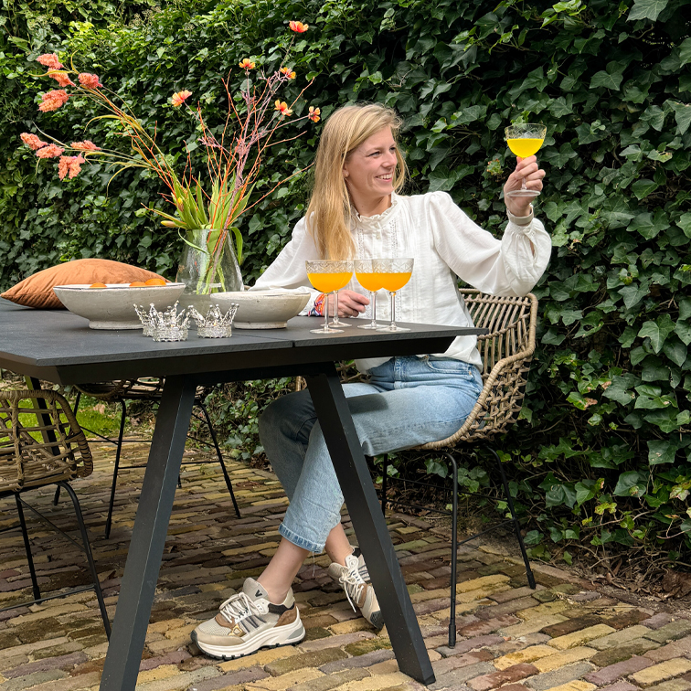 Femme en chemisier blanc et jean assise à une table noire extérieure, tenant un verre avec boisson orange, vase avec fleurs à côté