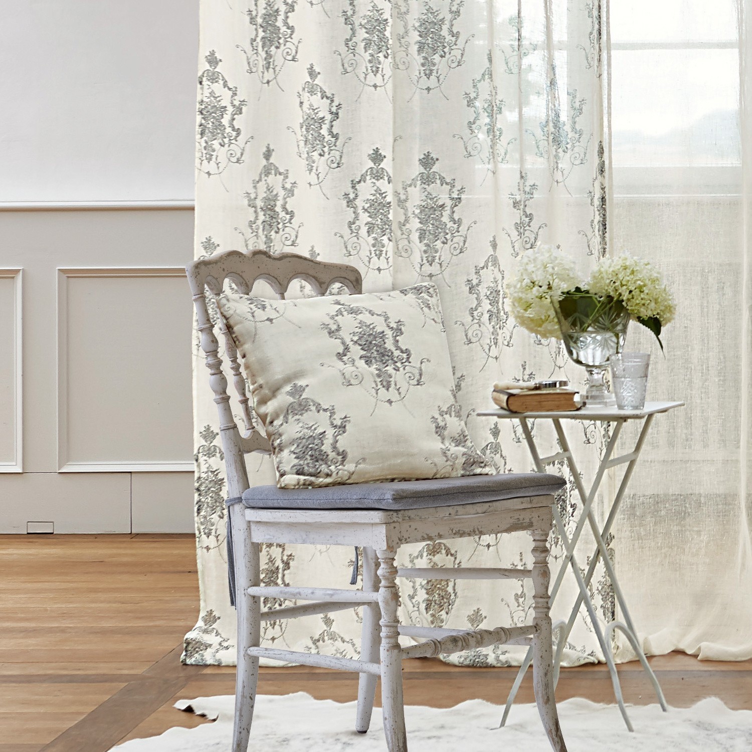 Housse de coussin Dinan Chaise en bois blanc avec coussin gris et coussin décoratif à motif floral, table avec fleurs à côté