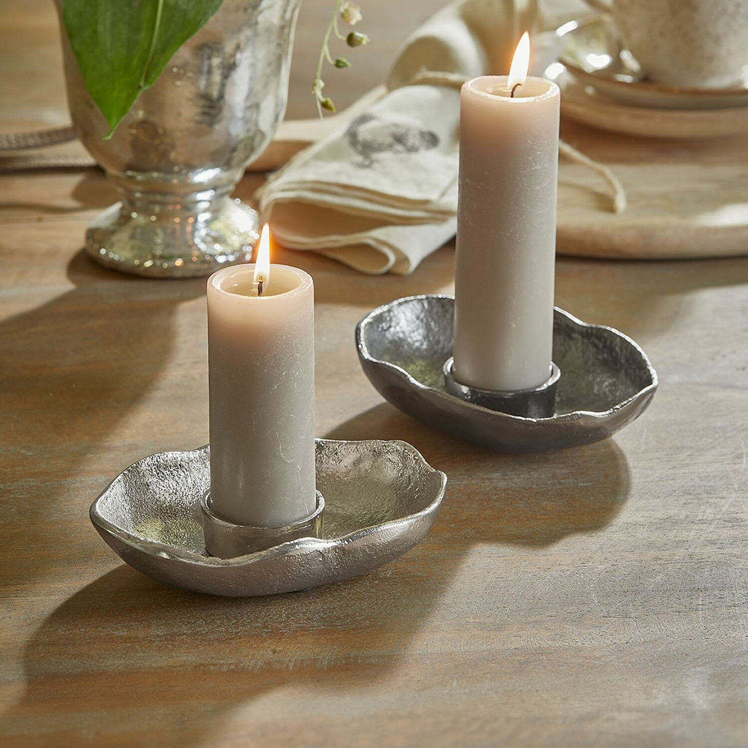 Lot de 2 porte-bougies chauffe-plats Quilvano Deux bougeoirs en métal argenté avec bougies grises sur une table en bois, décoration intérieure
