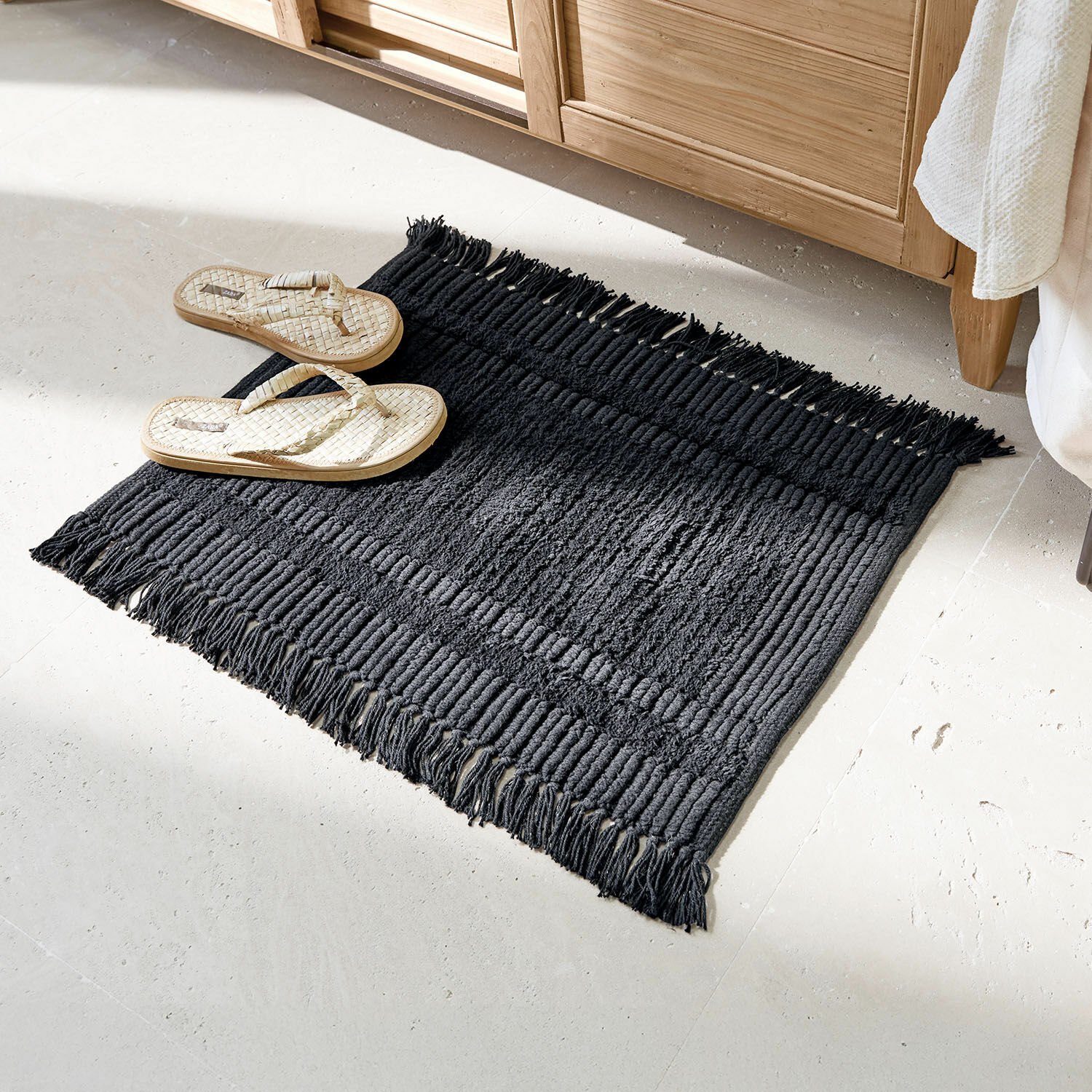 Tapis de bain Nexilo Tapis en coton noir avec franges sur sol clair, à côté des tongs tressées beiges