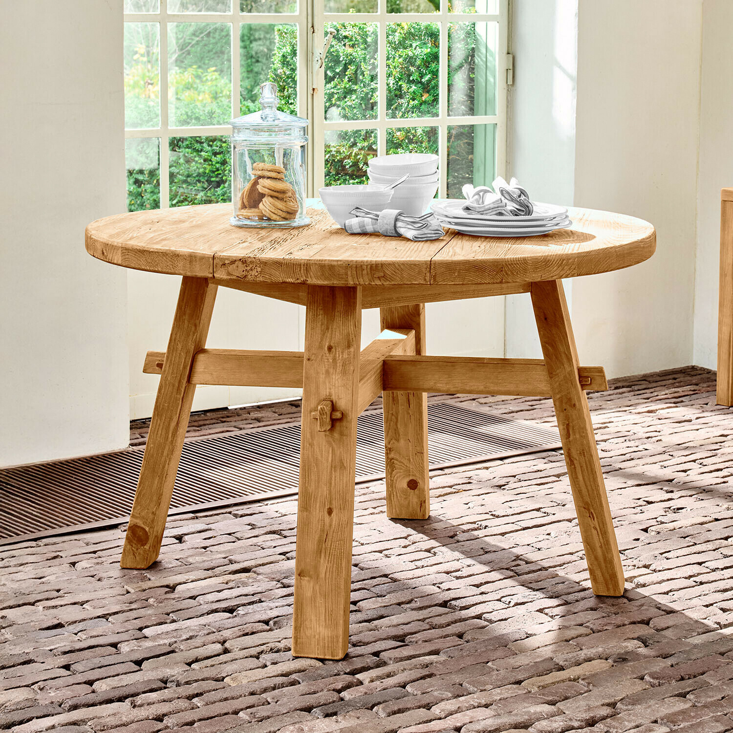 Table ronde en bois de pin naturel posée sur un sol en briques devant une fenêtre lumineuse
