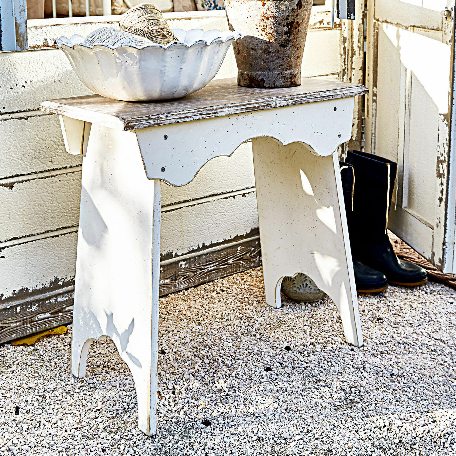 Tabouret Fria Table en bois blanc vieilli sur sol gravier avec bol décoratif et bottes noires à côté