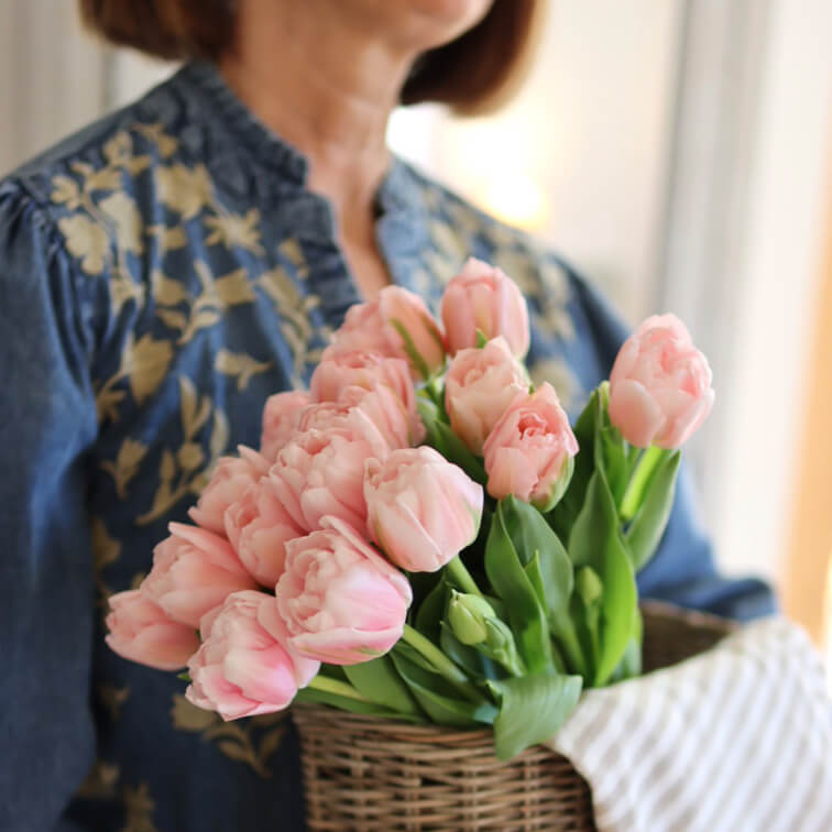 Bouquet de tulipes roses dans un panier en osier tenu par une femme en chemisier bleu à motif floral