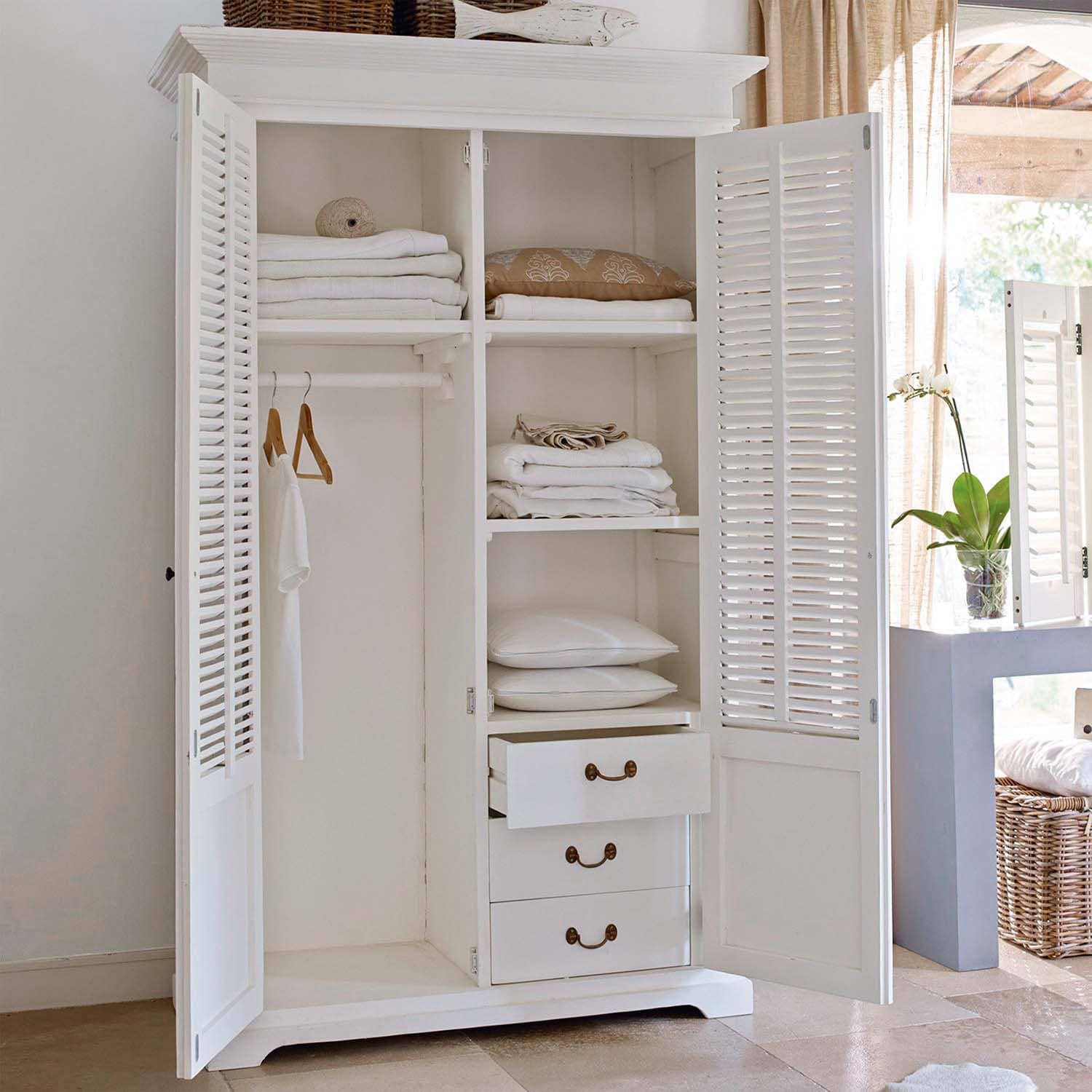 Armoire en bois blanc ouverte avec textiles blancs pliés et coussins dans une pièce lumineuse