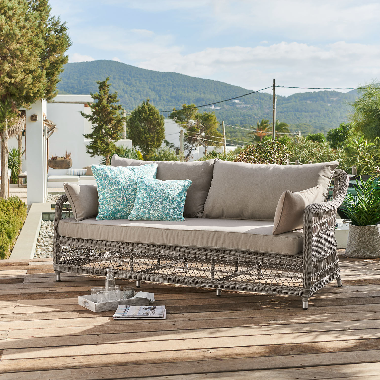 Canapé Tanaiste Canapé d'extérieur en rotin gris avec coussins beige et coussins décoratifs turquoise sur terrasse en bois