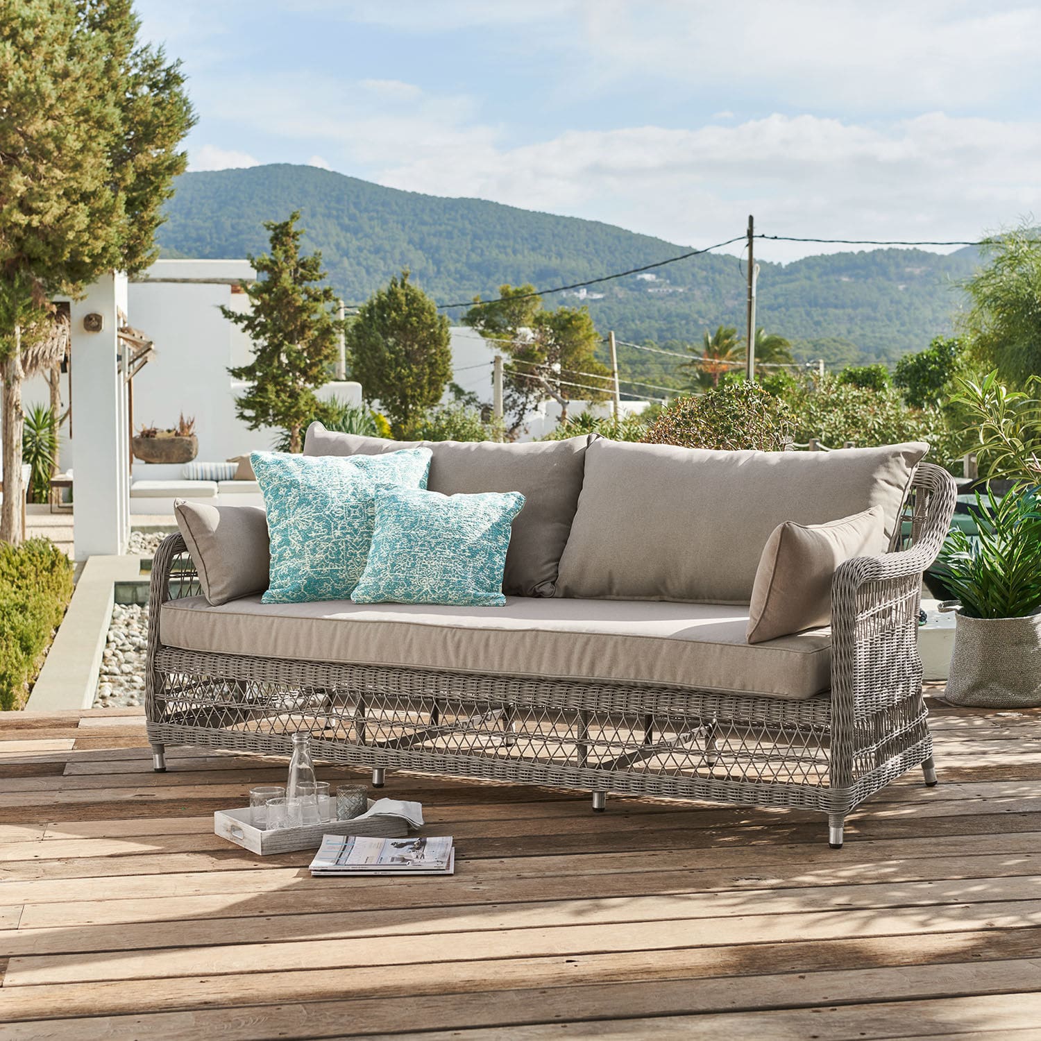 Canapé Tanaiste Canapé d'extérieur en rotin gris avec coussins beige et coussins décoratifs turquoise sur terrasse en bois