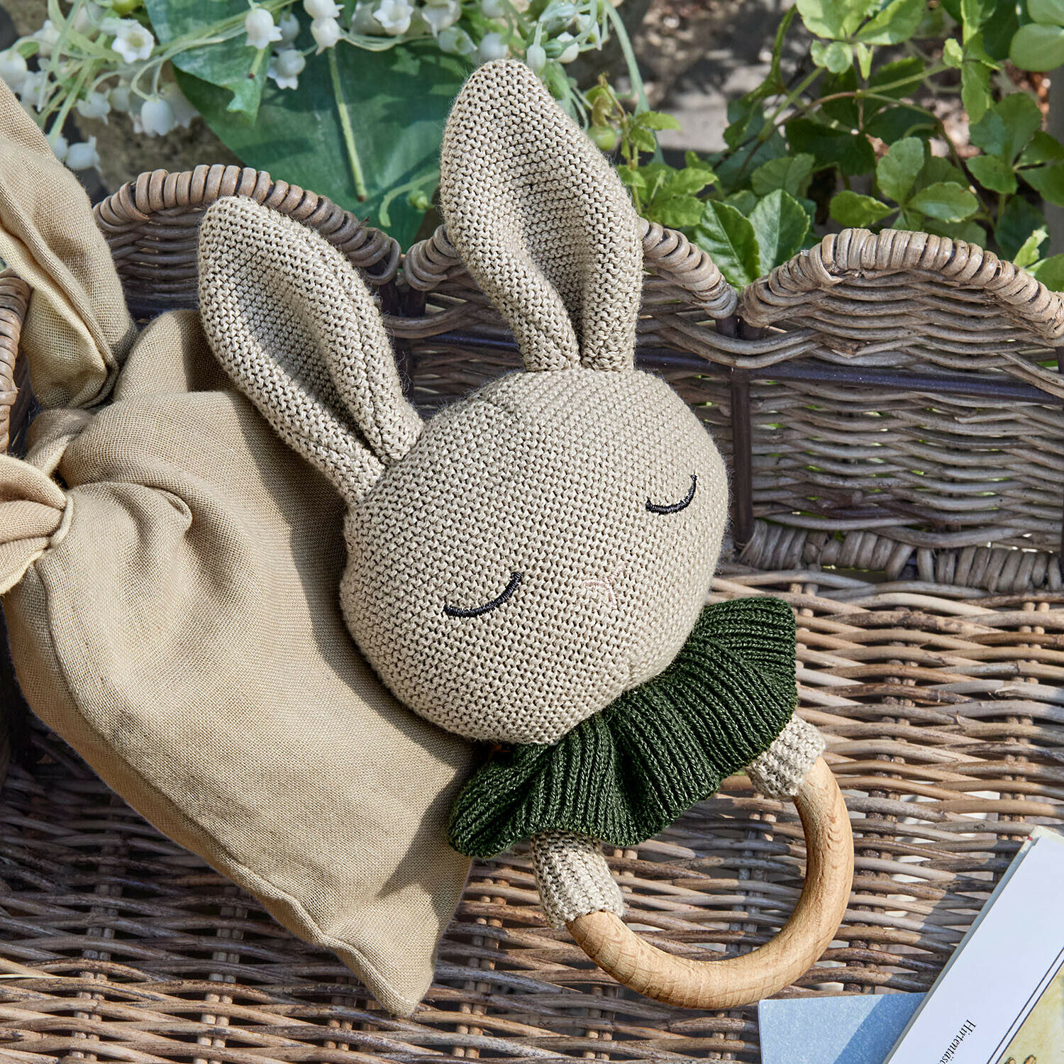 Anneau de jouets Cuddlecloud Hochet lapin en tricot beige avec col vert et anneau en bois sur fond en osier
