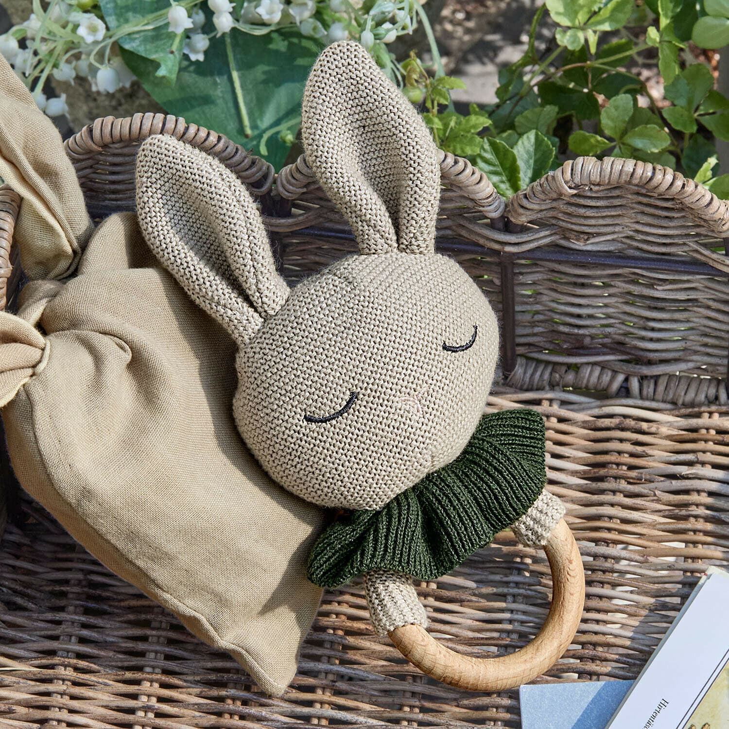 Anneau de jouets Cuddlecloud Hochet lapin en tricot beige avec col vert et anneau en bois sur fond en osier